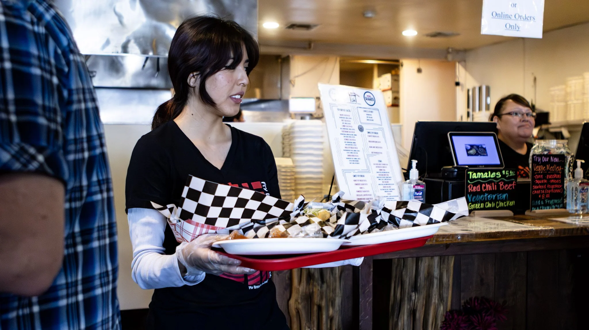 A woman serving food at a restaurant counter, holding a tray with wrapped food items, next to a man in a plaid shirt; behind the counter are signs, a sanitizer, and a woman facing the camera.