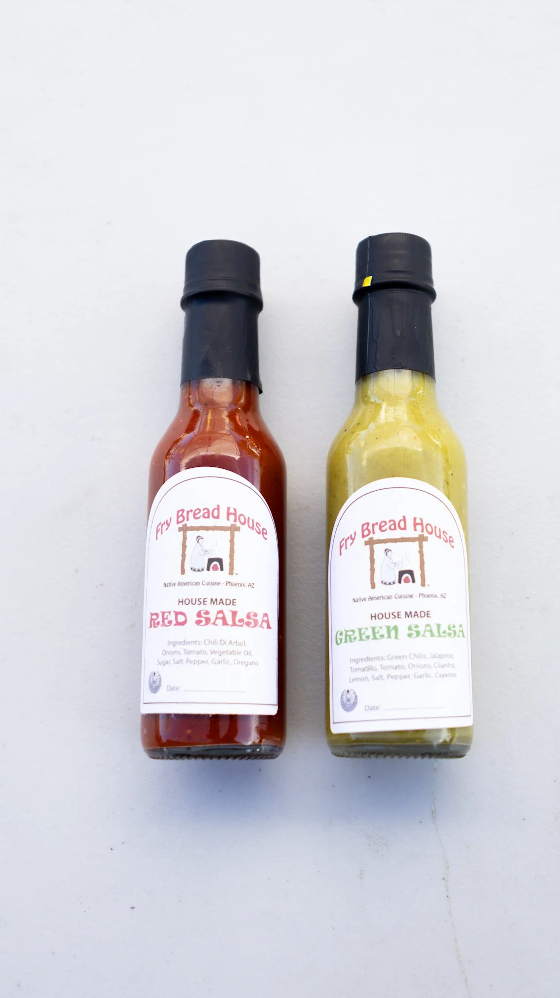 Two bottles of homemade salsa, one red and one green, from Fry Bread House in Phoenix, Arizona, placed on a white surface.