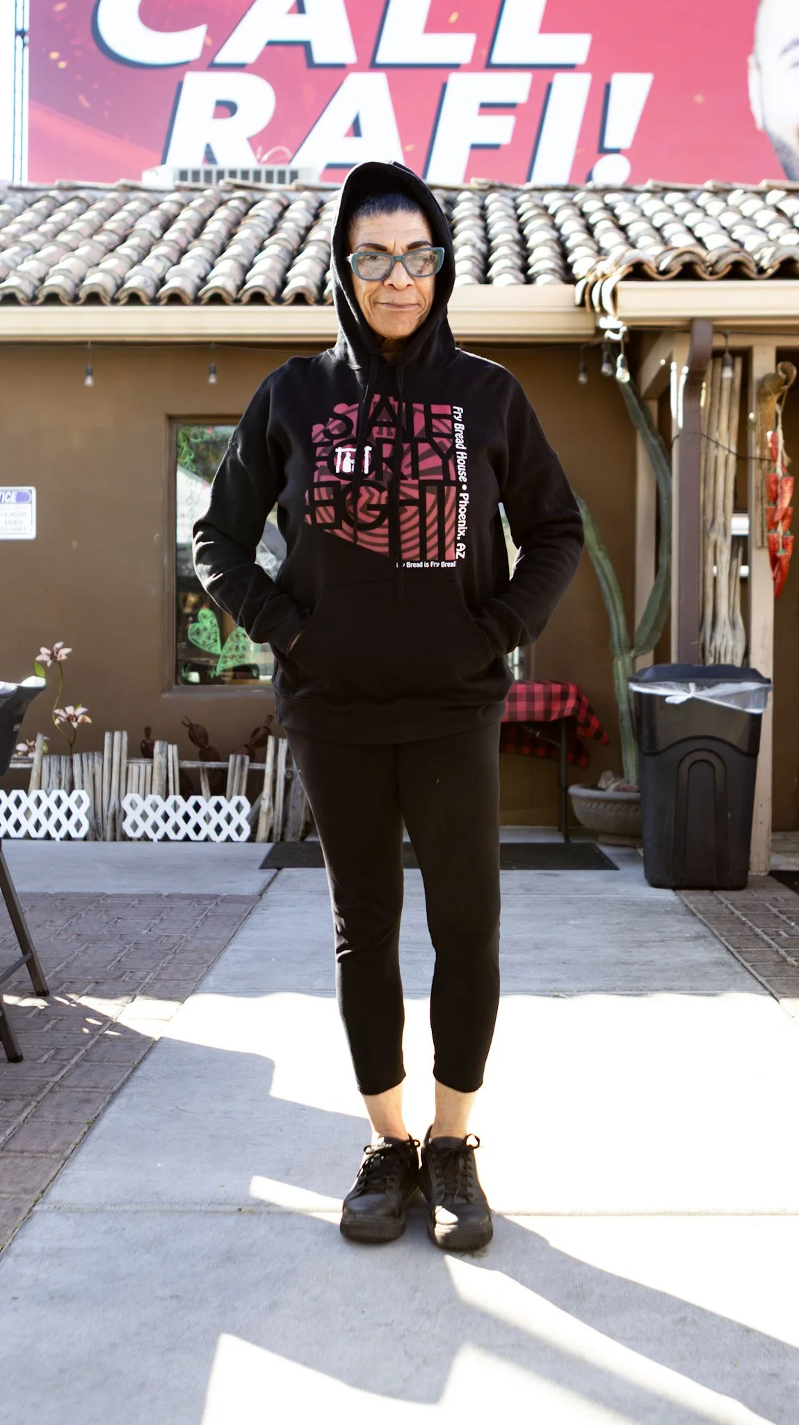 A woman in a black hoodie and black pants standing outside in front of a restaurant with a large pink sign that says 'CALL RAFE!'