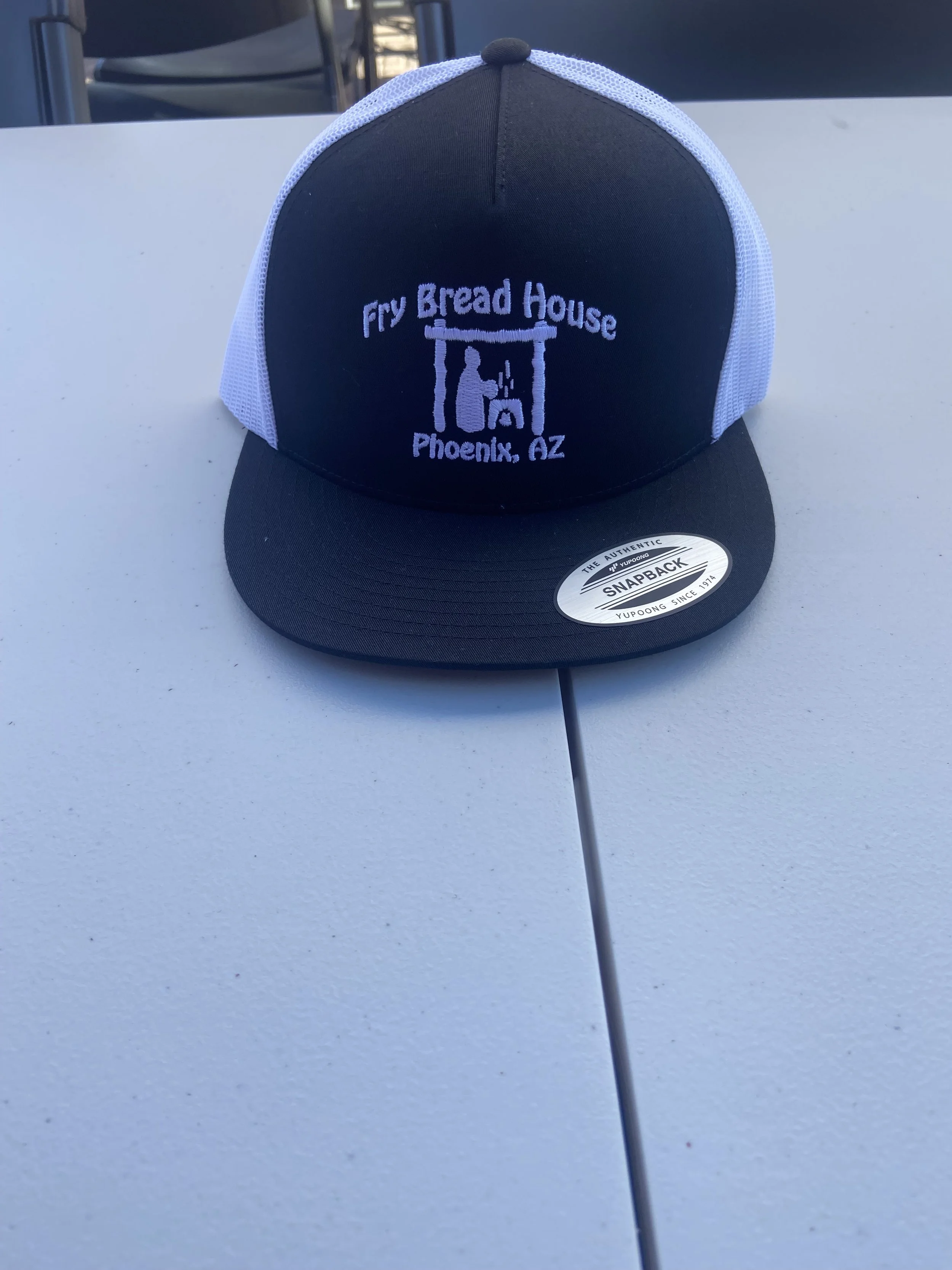 A black and white trucker hat with embroidery that reads 'Fry Bread House, Phoenix, AZ' and a graphic of a person under a structure, on a white surface.