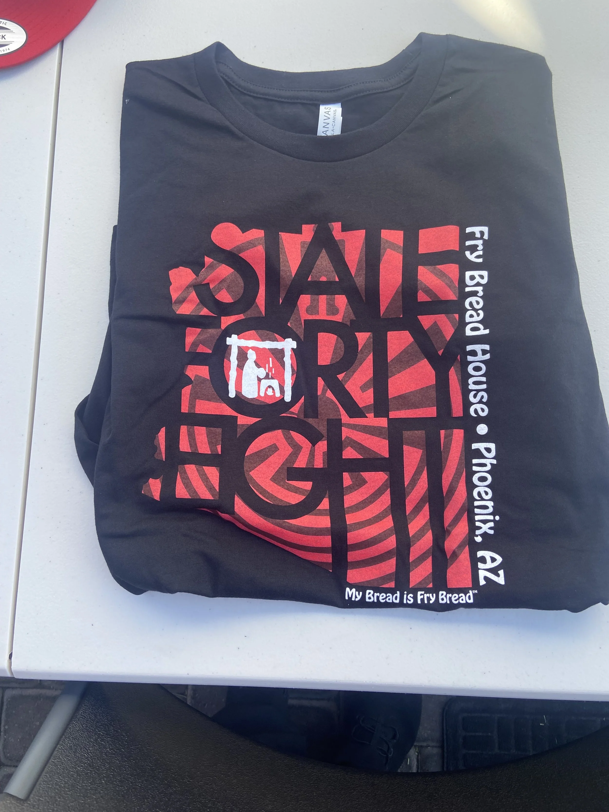 Folded black t-shirt with a red circular design and the text 'State of Fry' in large black letters, along with smaller text 'Fry Bread House Phoenix AZ' and 'My Bread is Fry Bread'.