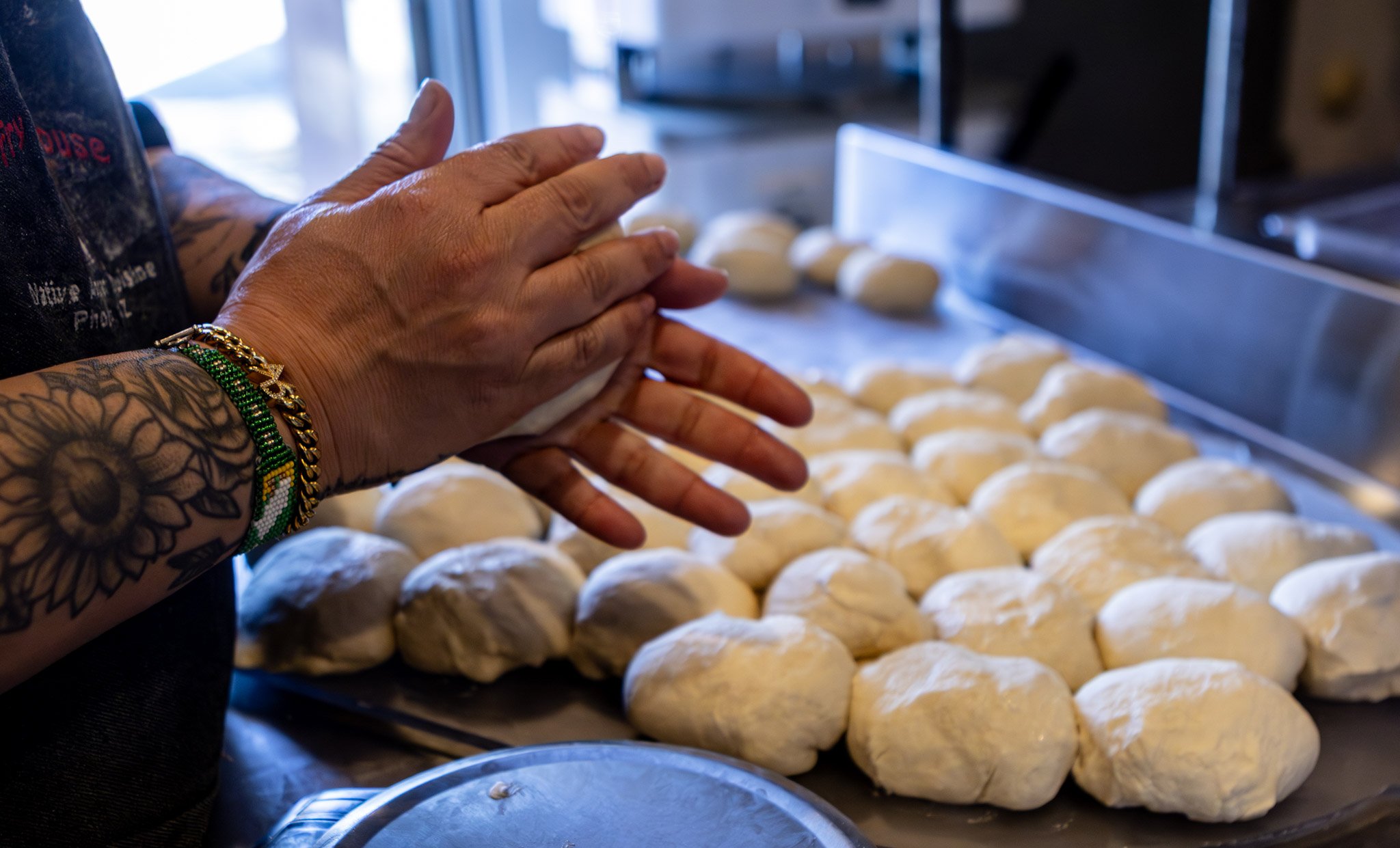 Person with tattooed hands and bracelets shaping dough into balls on a tray in a bakery or kitchen.