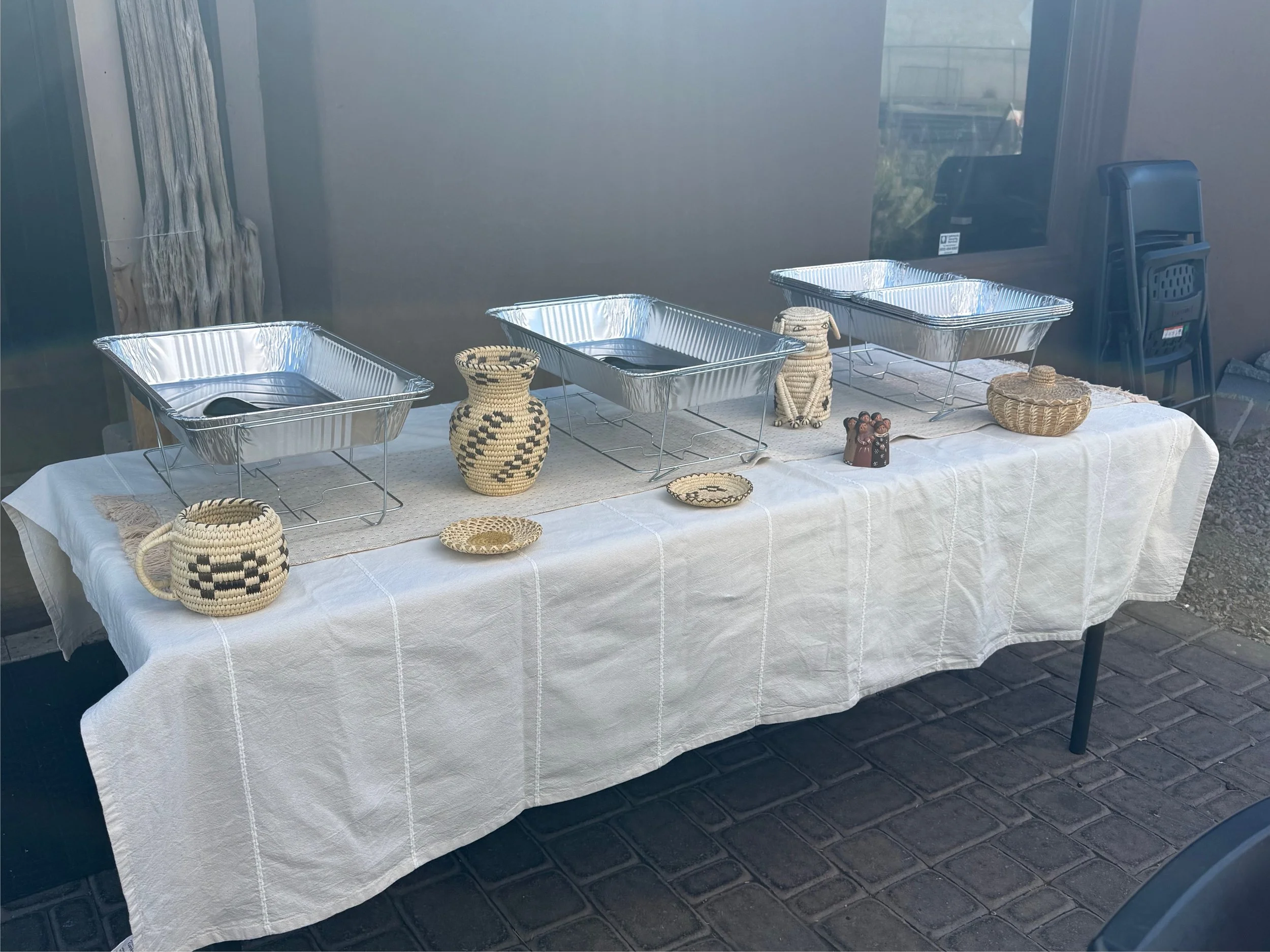 Table covered with a white cloth displaying three aluminum chafing dishes, various woven ceramic and basket items, and small decorative items, set against a glass wall, with chairs and outdoor scenery in the background.