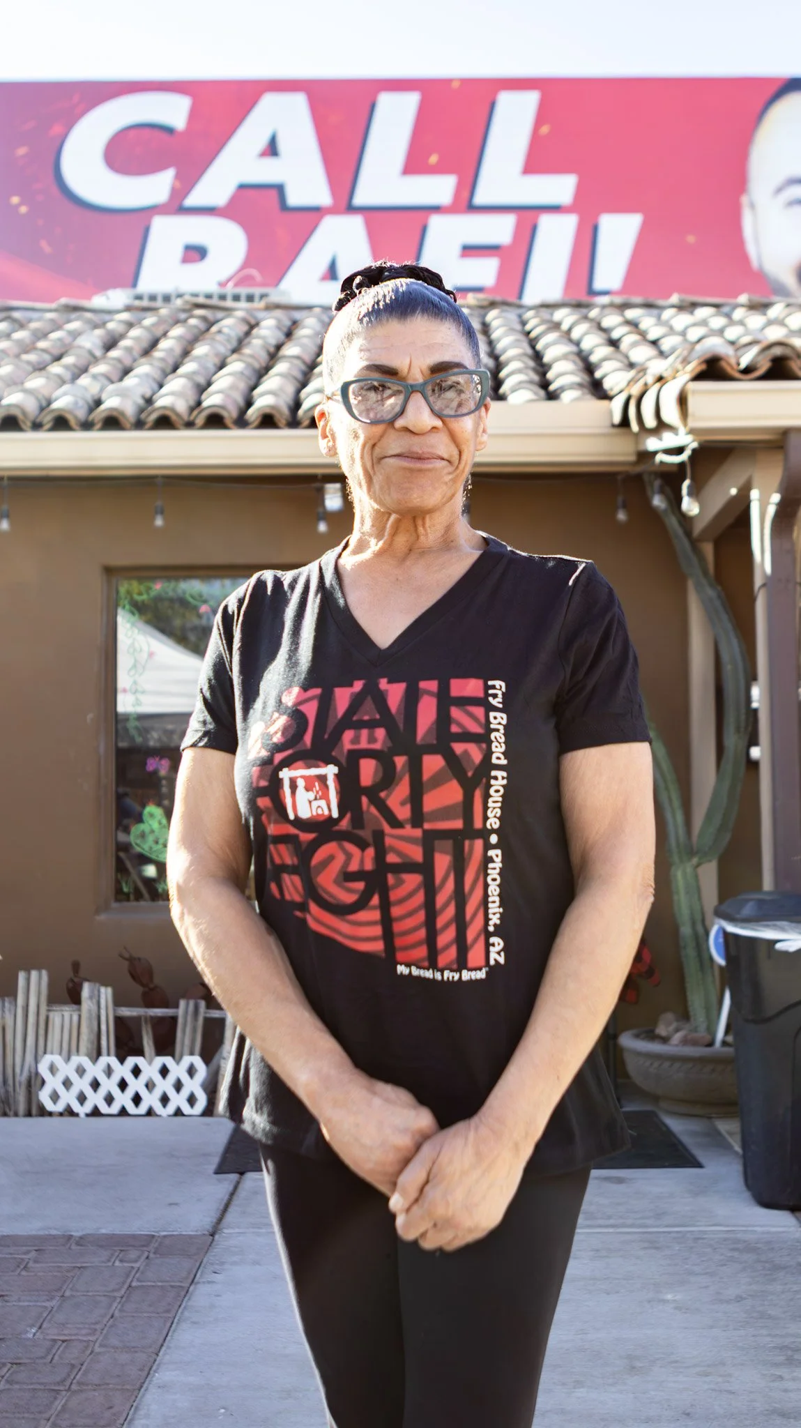 A woman standing outside in front of a building with a large red and white sign that says 'Call Ball.' She is wearing a black T-shirt with red and white graphics and text, including 'State Fair' and 'My bread is Fry Bread.' She has dark hair pulled b