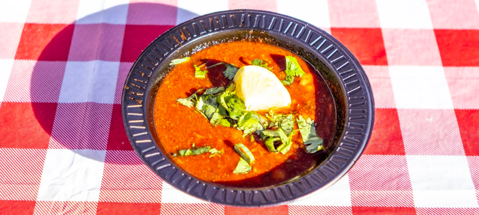 A bowl of Indian red-colored curry garnished with cilantro, a lemon wedge, and herbs, placed on a red and white checkered tablecloth.