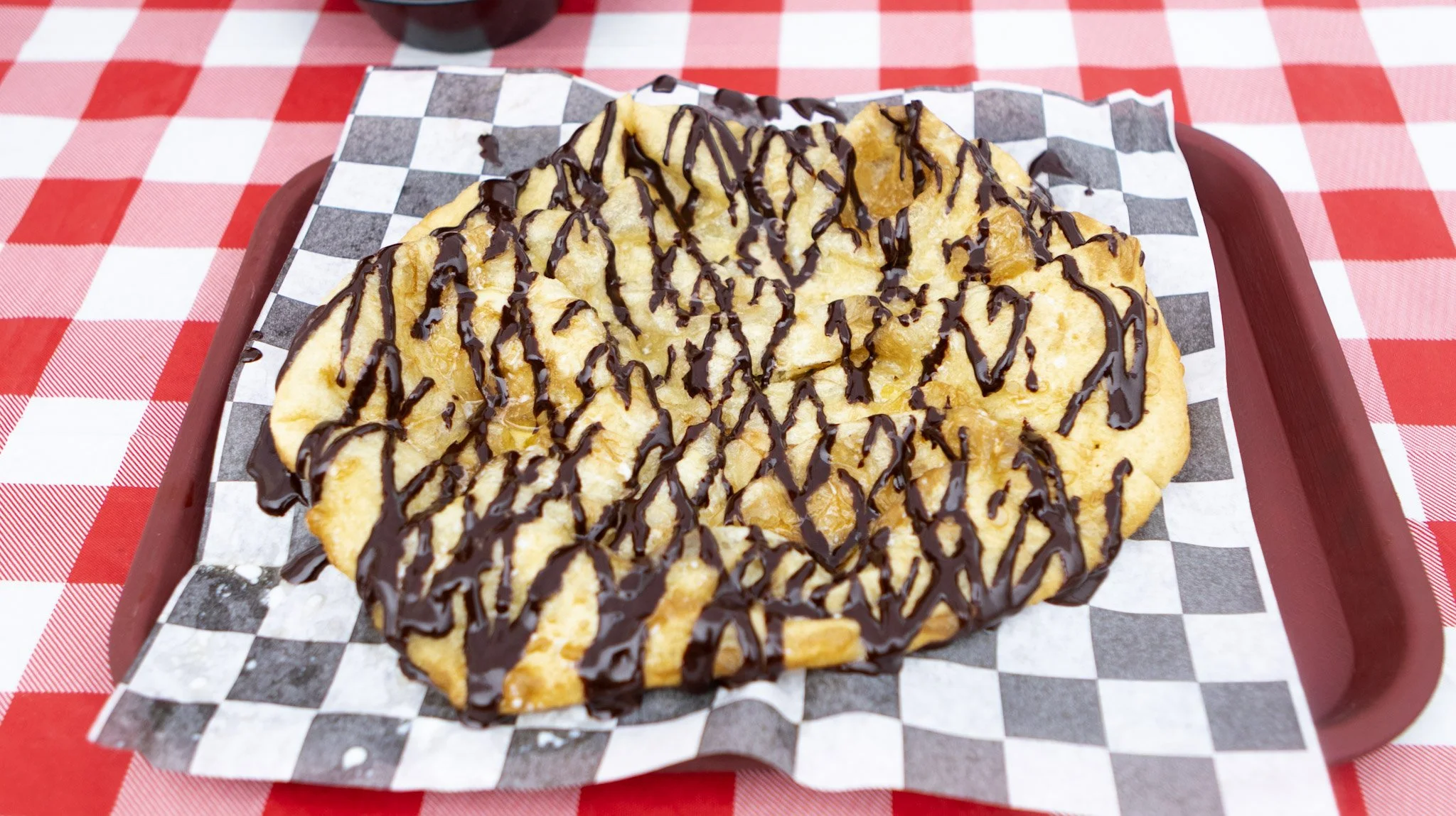 Fried dough topped with chocolate syrup on checkered paper, served on a red tray, with a red and white checkered tablecloth.
