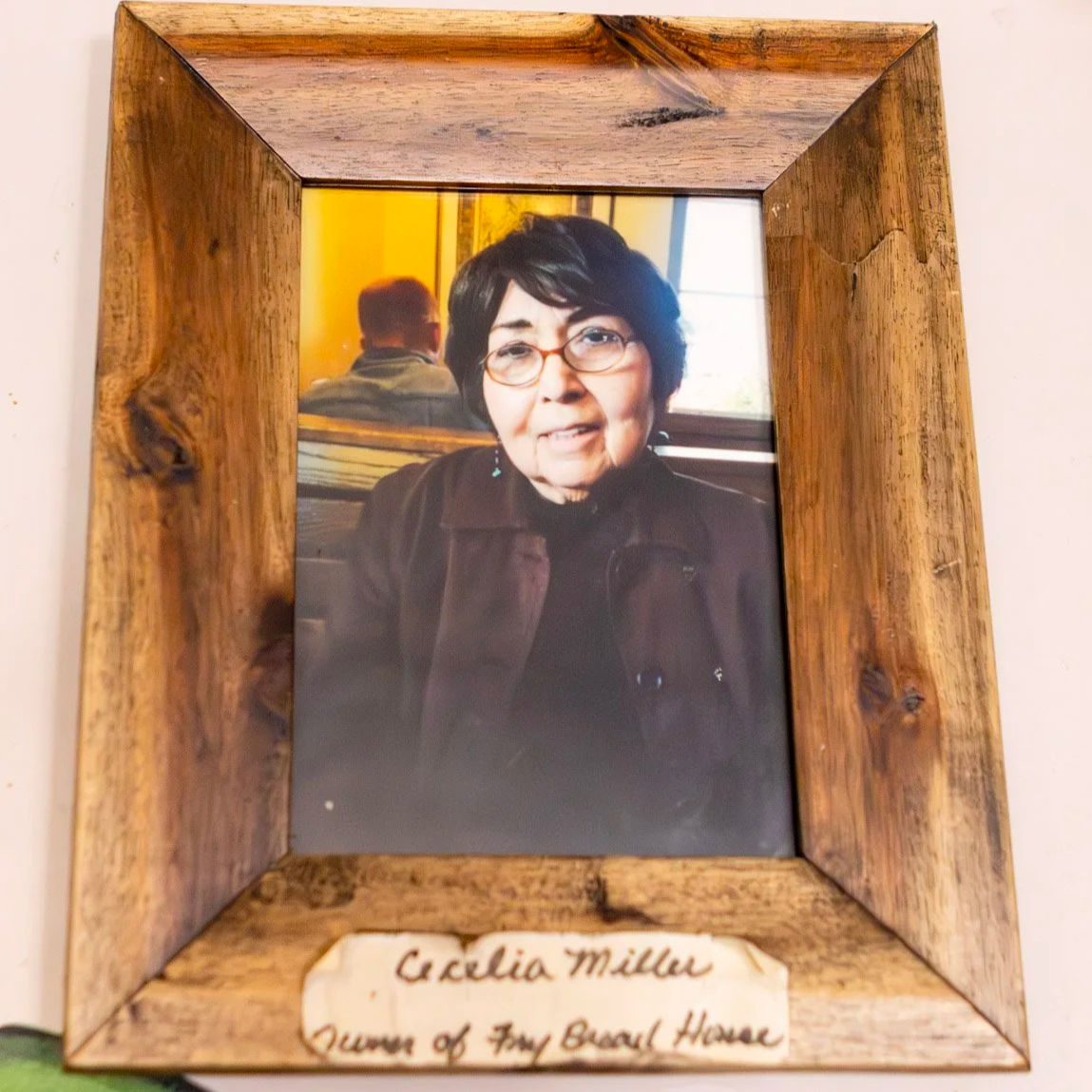 Framed photograph of an elderly woman with black hair, glasses, and earrings, sitting indoors near a window, with a man with glasses and a light-colored shirt visible in the background. The frame is made of rustic wood, and there is a handwritten note at the bottom that reads, 'Cecilia Miller, Queen of Fry Bread House.'