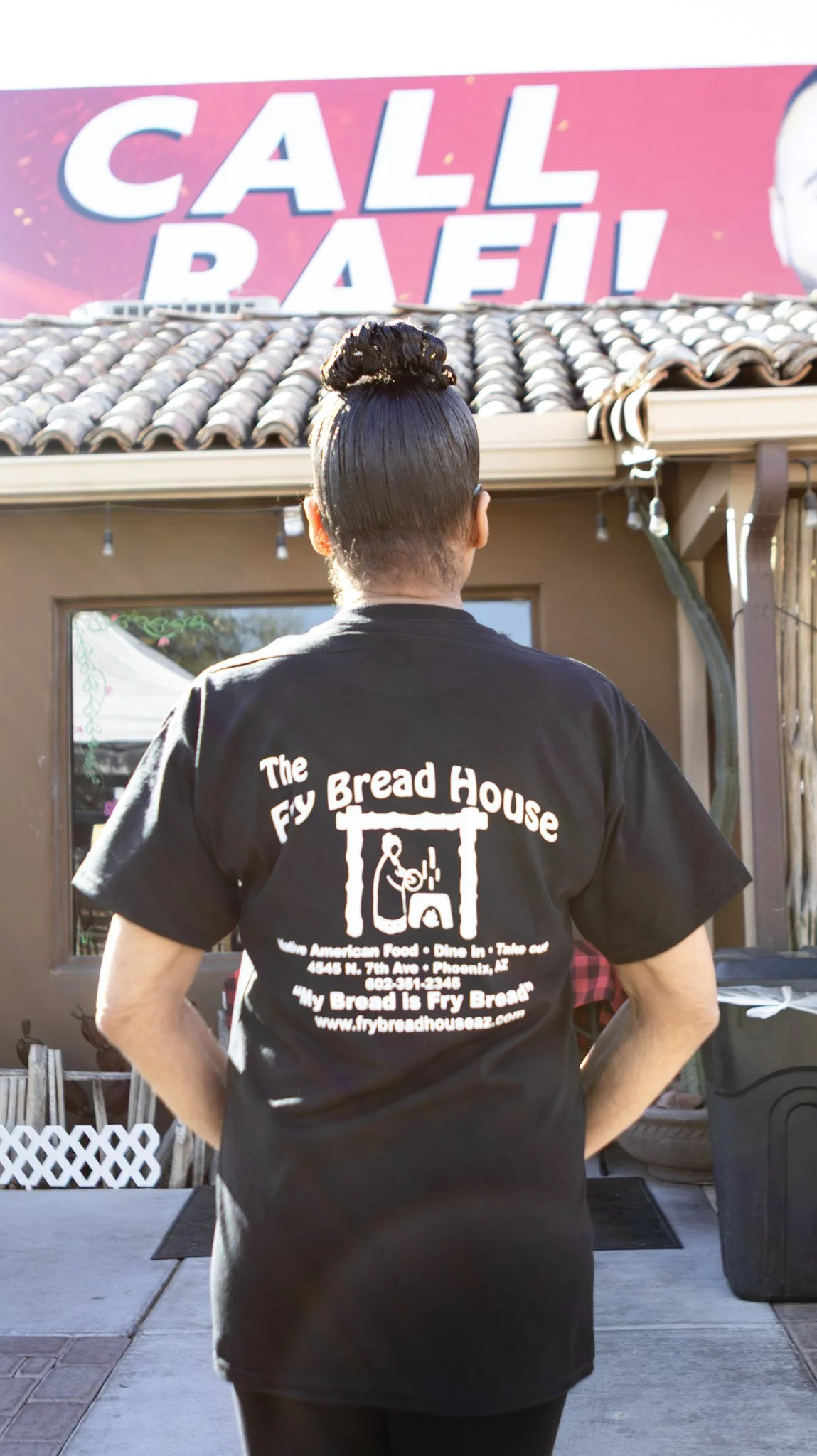 A person with dark hair styled in a bun, wearing a black t-shirt with the logo and name of The Fry Bread House on the back, standing outside a building with a sign that says 'CALL.'