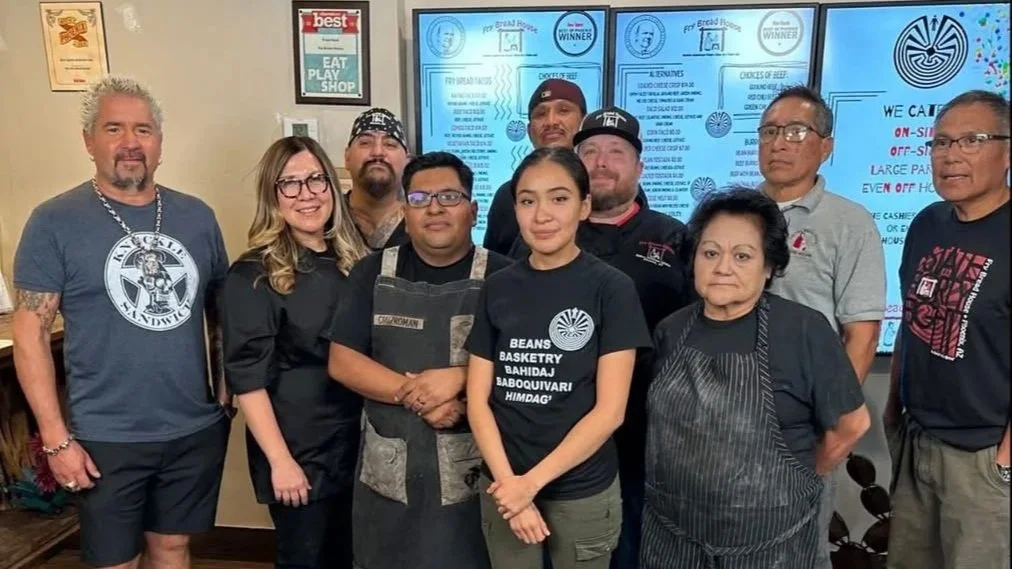 A diverse group of ten people, including men and women, posing together indoors. Some are wearing casual clothes, and one woman is wearing an apron. They are standing in front of a digital menu display with various restaurant options and awards, indicating it is a restaurant setting.