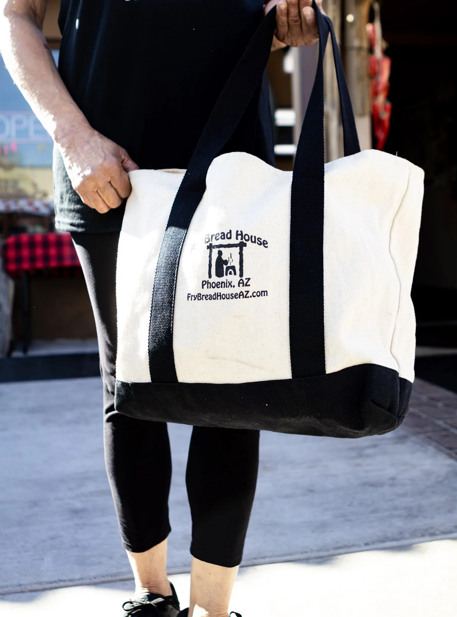 Person carrying a beige tote bag with black straps, featuring the logo and text for Bread House in Phoenix, AZ.