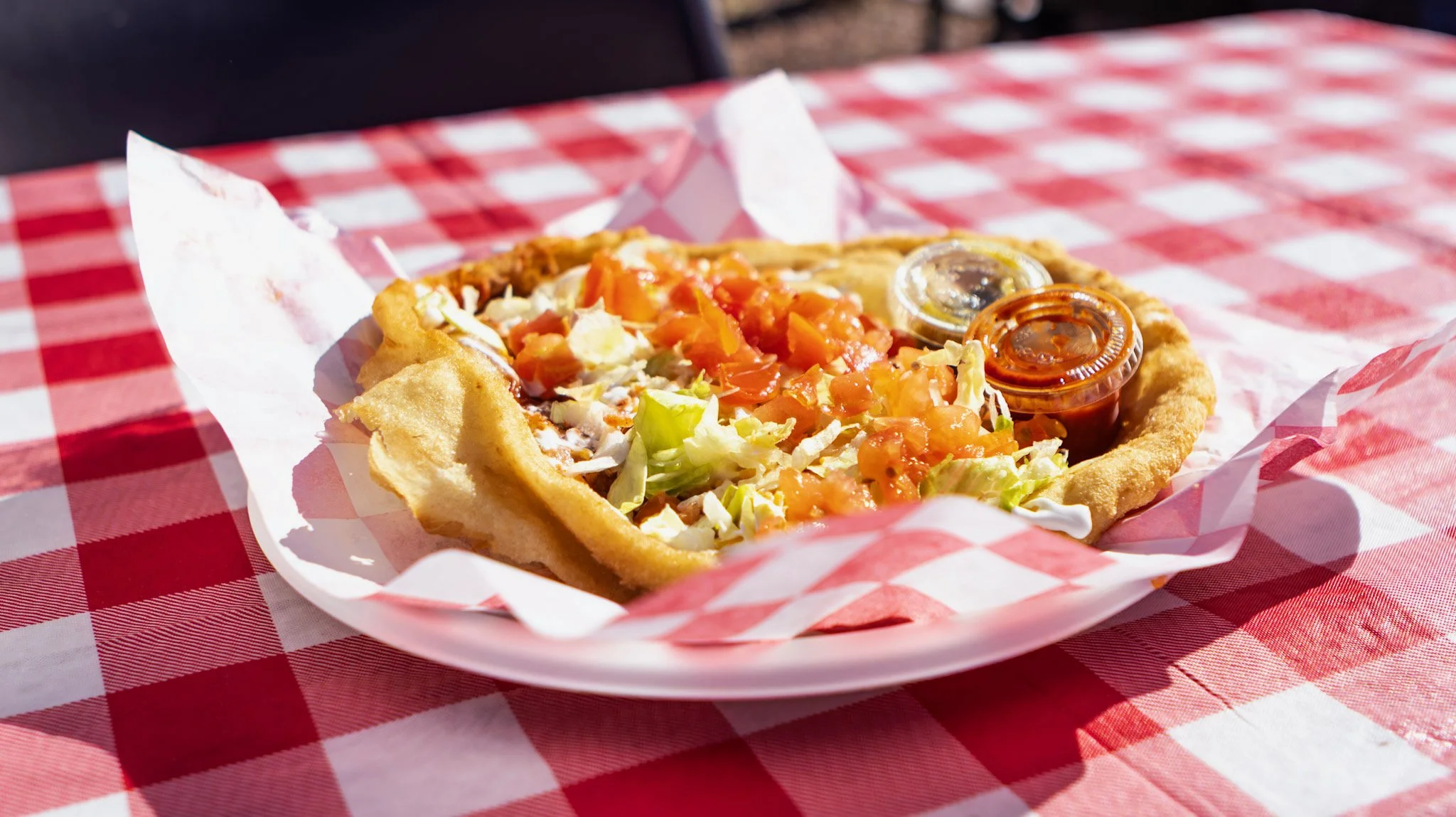 Deep-fried taco with chopped lettuce, tomatoes, shredded cheese, and two small containers of sauce on a red and white checkered tablecloth.