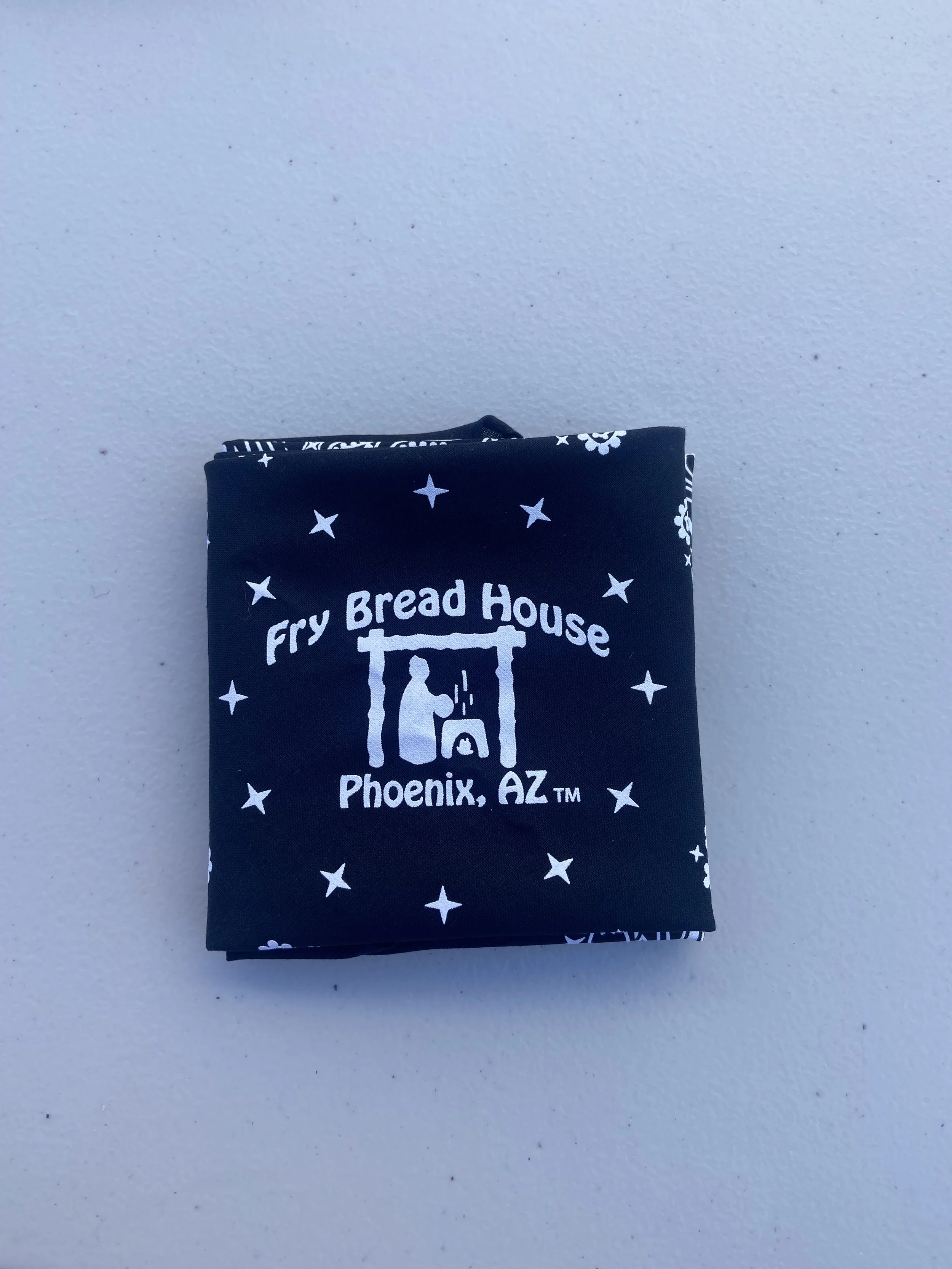 A black cloth with white text and design, reading 'Fry Bread House Phoenix, AZ,' featuring a graphic of a person cooking over a fire.