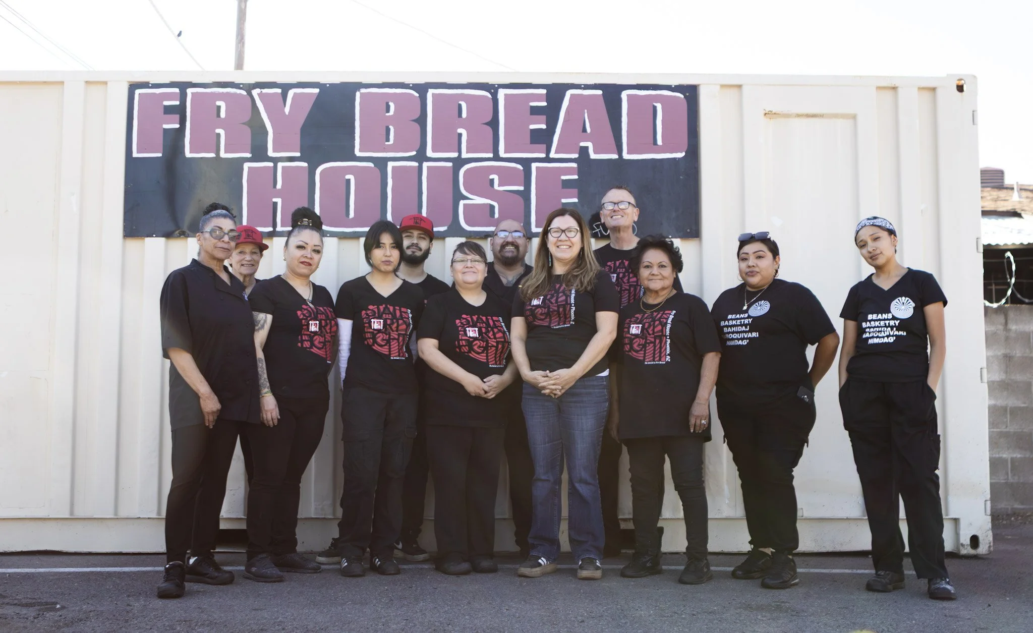 Group of ten people standing in front of a sign that reads 'Fry Bread House.' They are dressed casually, some wearing black t-shirts with a red and black design, and they are smiling at the camera.
