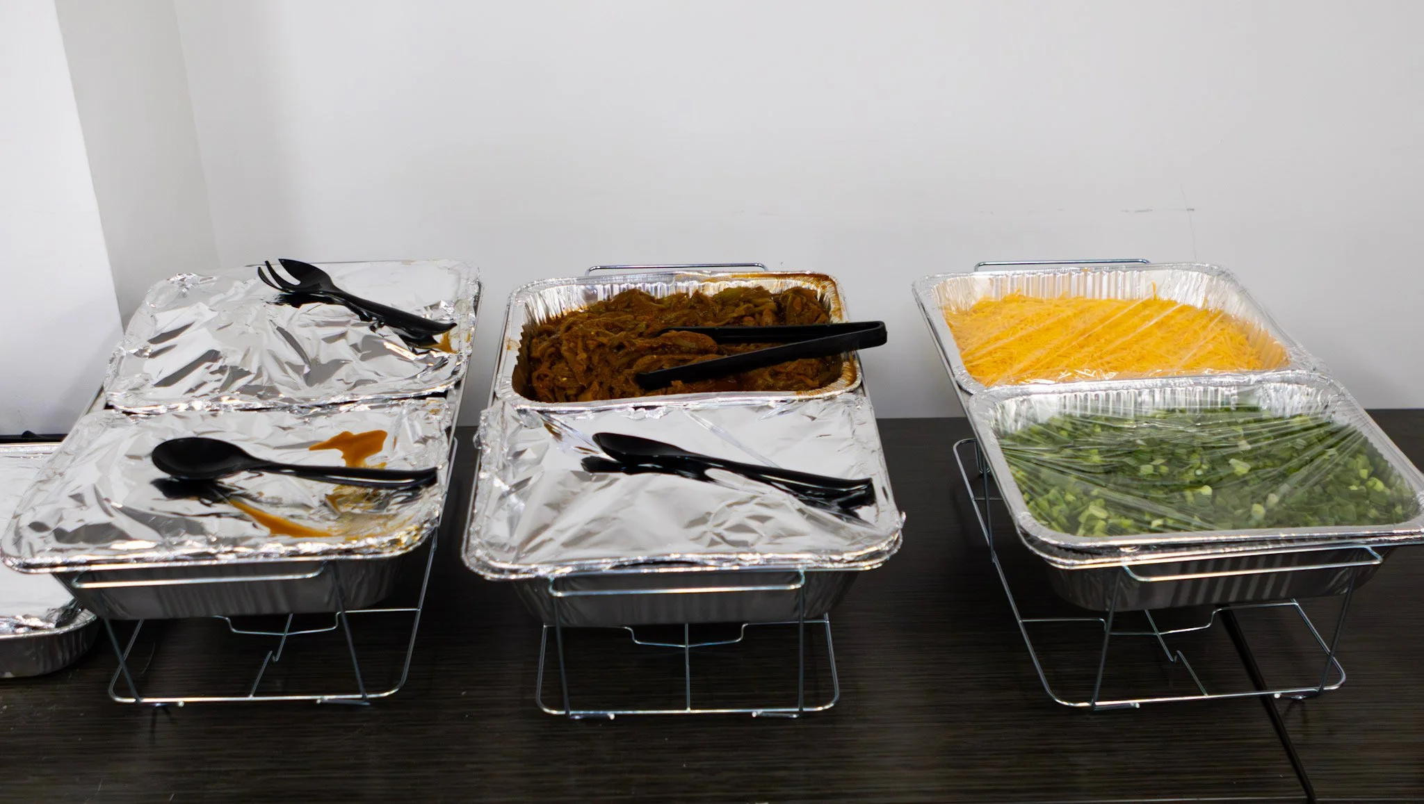 Buffet of pre-arranged food trays with aluminum foil covers, some uncovered showing noodles, shredded cheese, and chopped greens, on a dark wooden table against a white wall.