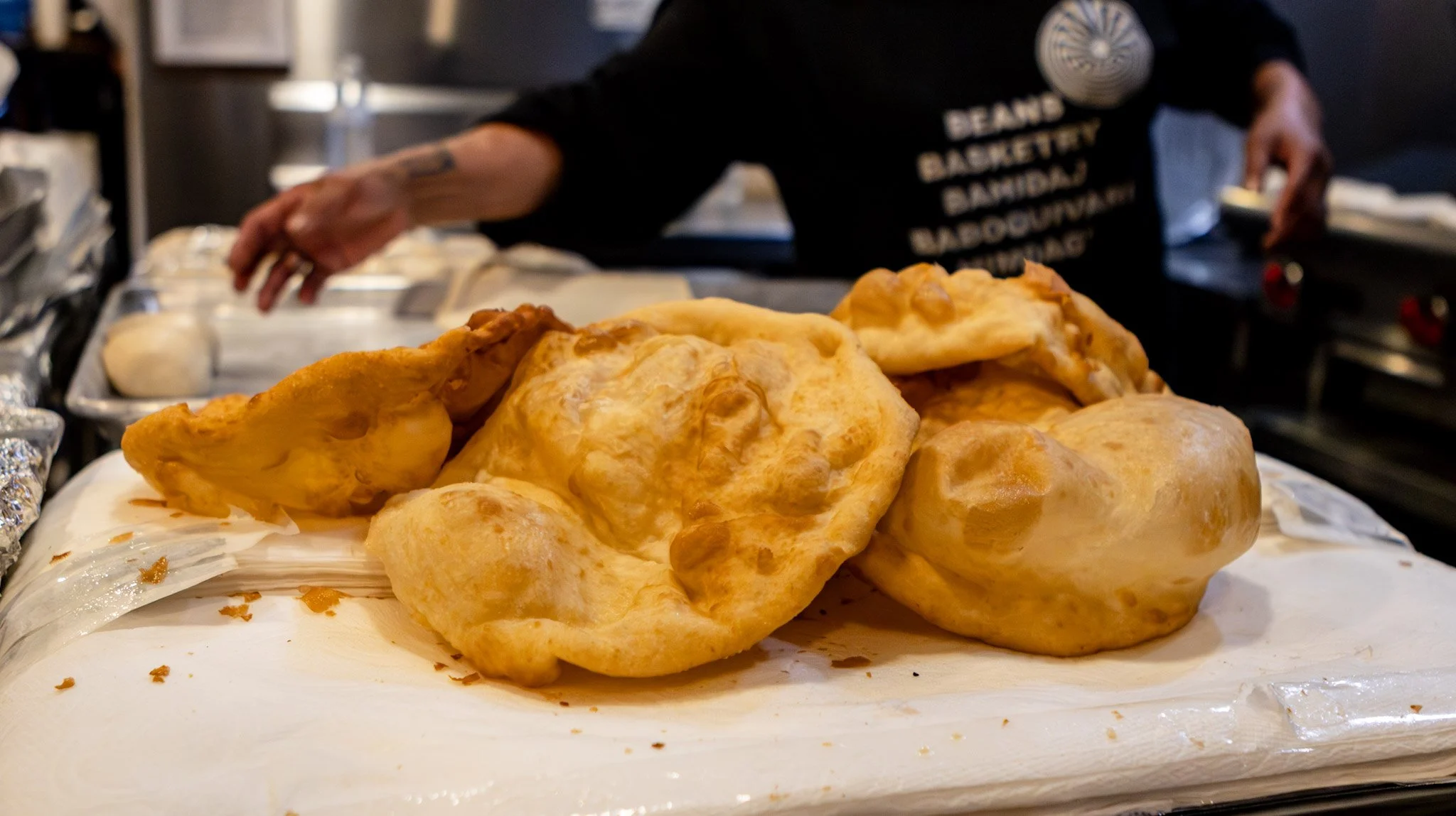 Freshly made, golden brown, fried Indian bread called 'bhatura' placed on a white surface with people preparing food in the background.