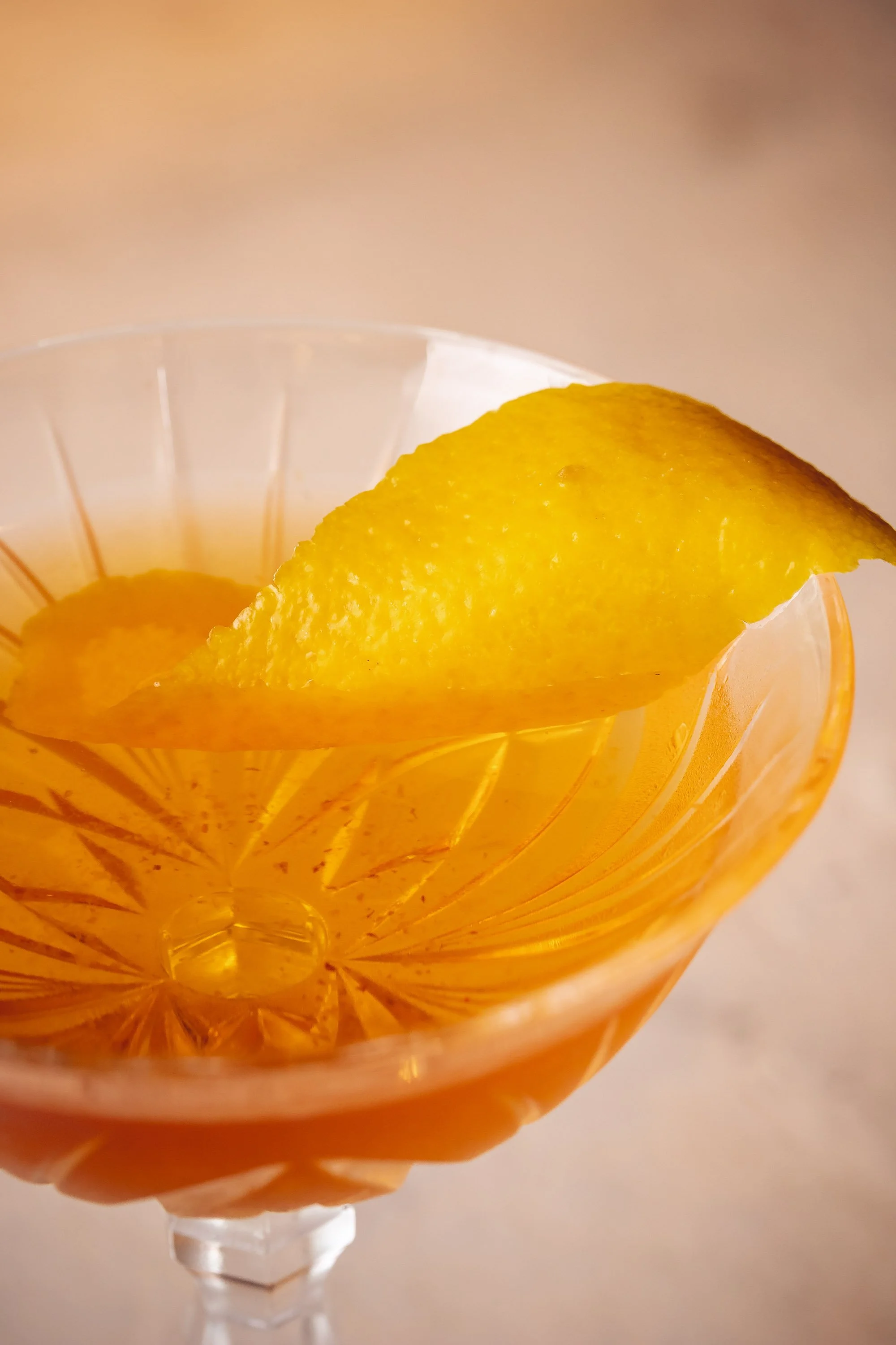 Orange cocktail in coupe glass with lemon peel garnish against soft neutral background
