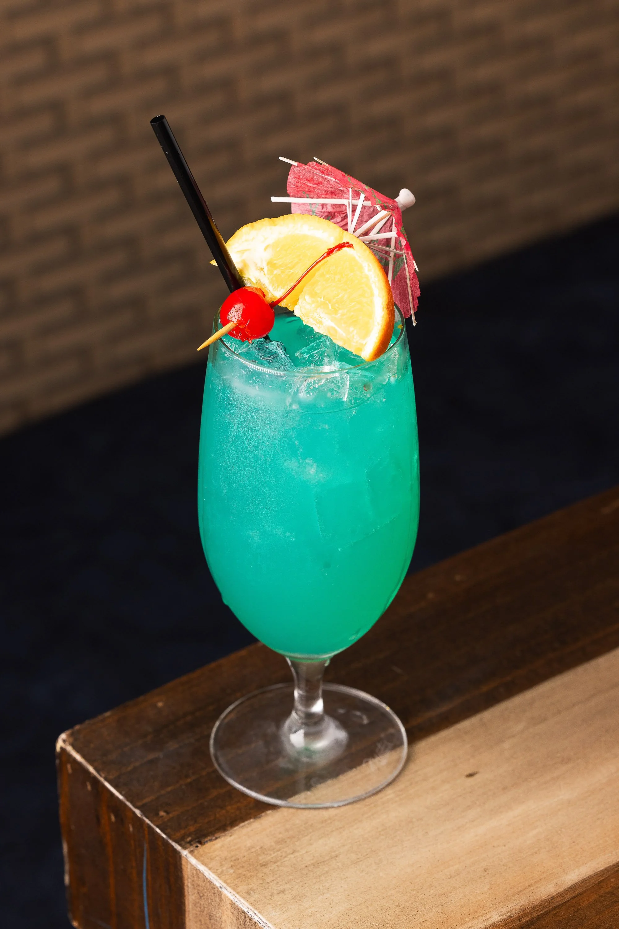 Blue tropical cocktail with orange slice, cherry, and paper umbrella in tall stemmed glass
