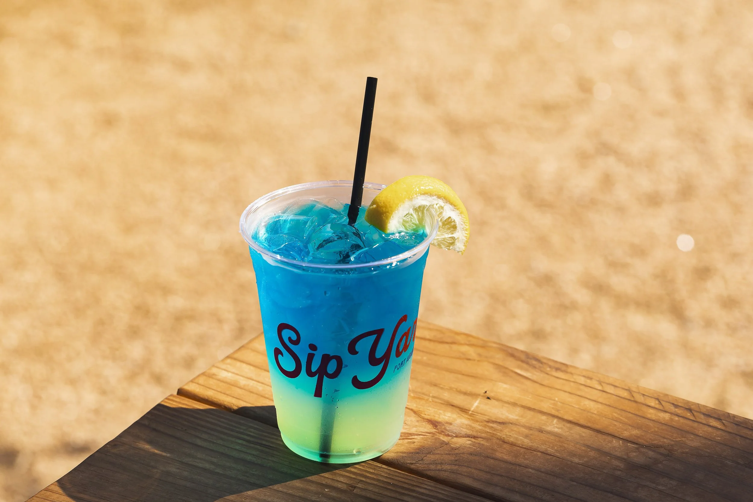 Blue layered frozen cocktail in branded plastic cup with lemon wedge on outdoor wooden table
