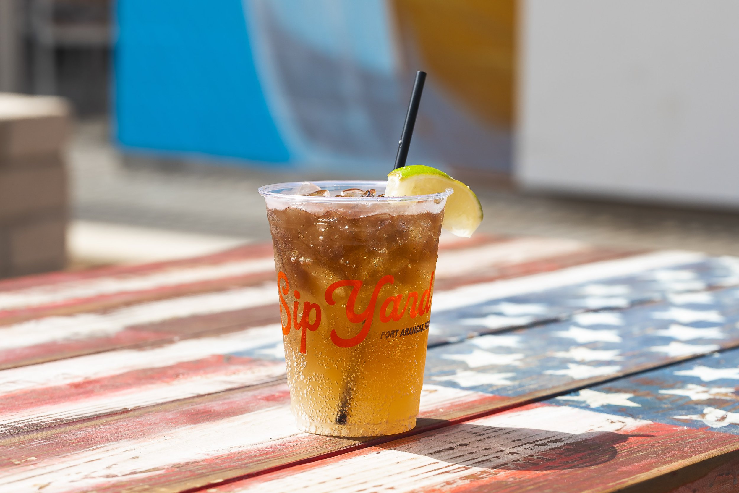 Iced amber cocktail in branded plastic cup with lime wedge on weathered painted outdoor table
