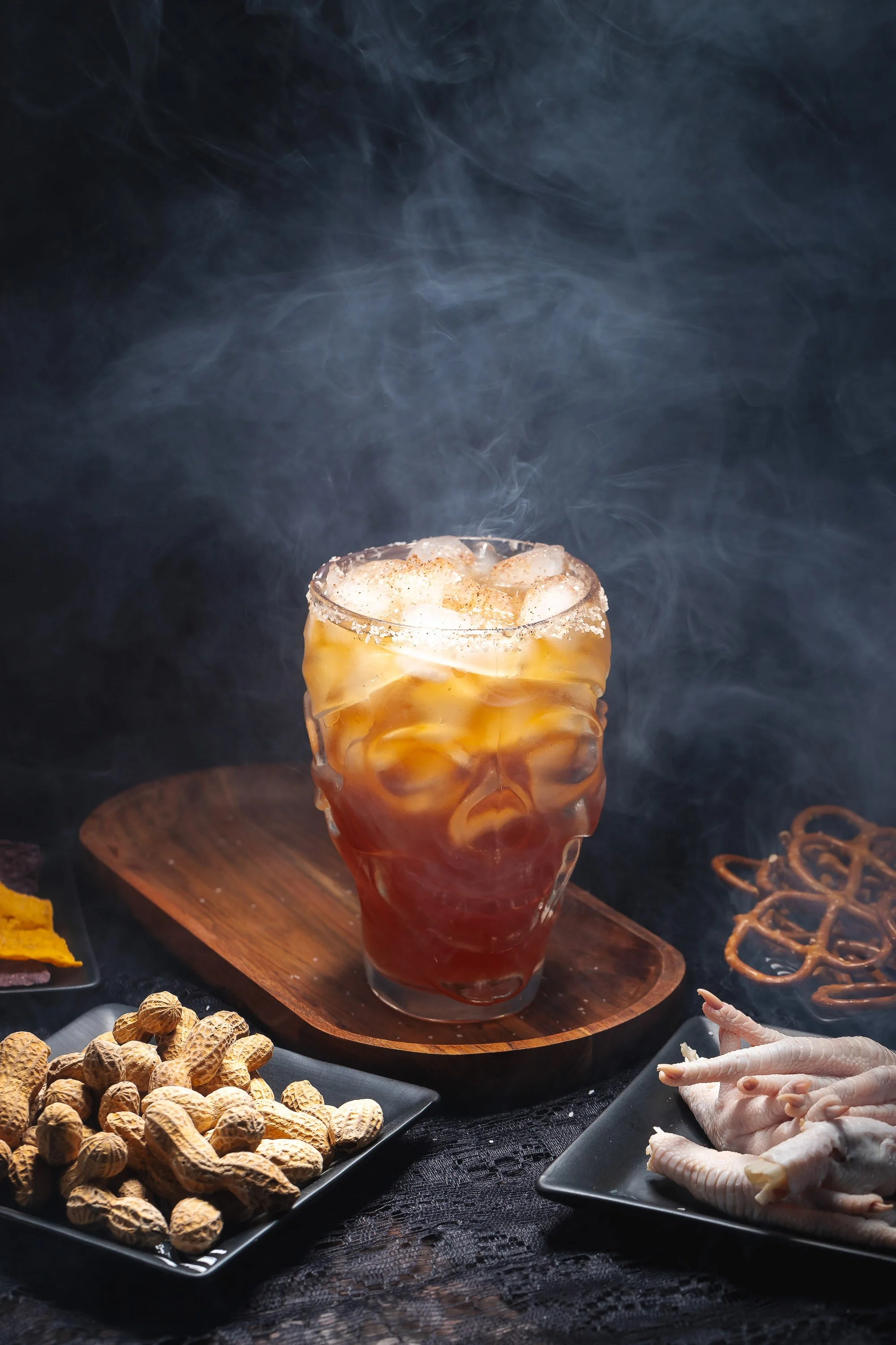 Halloween-themed smoked cocktail in skull glass with dramatic fog, dark moody styling, and spooky snack props
