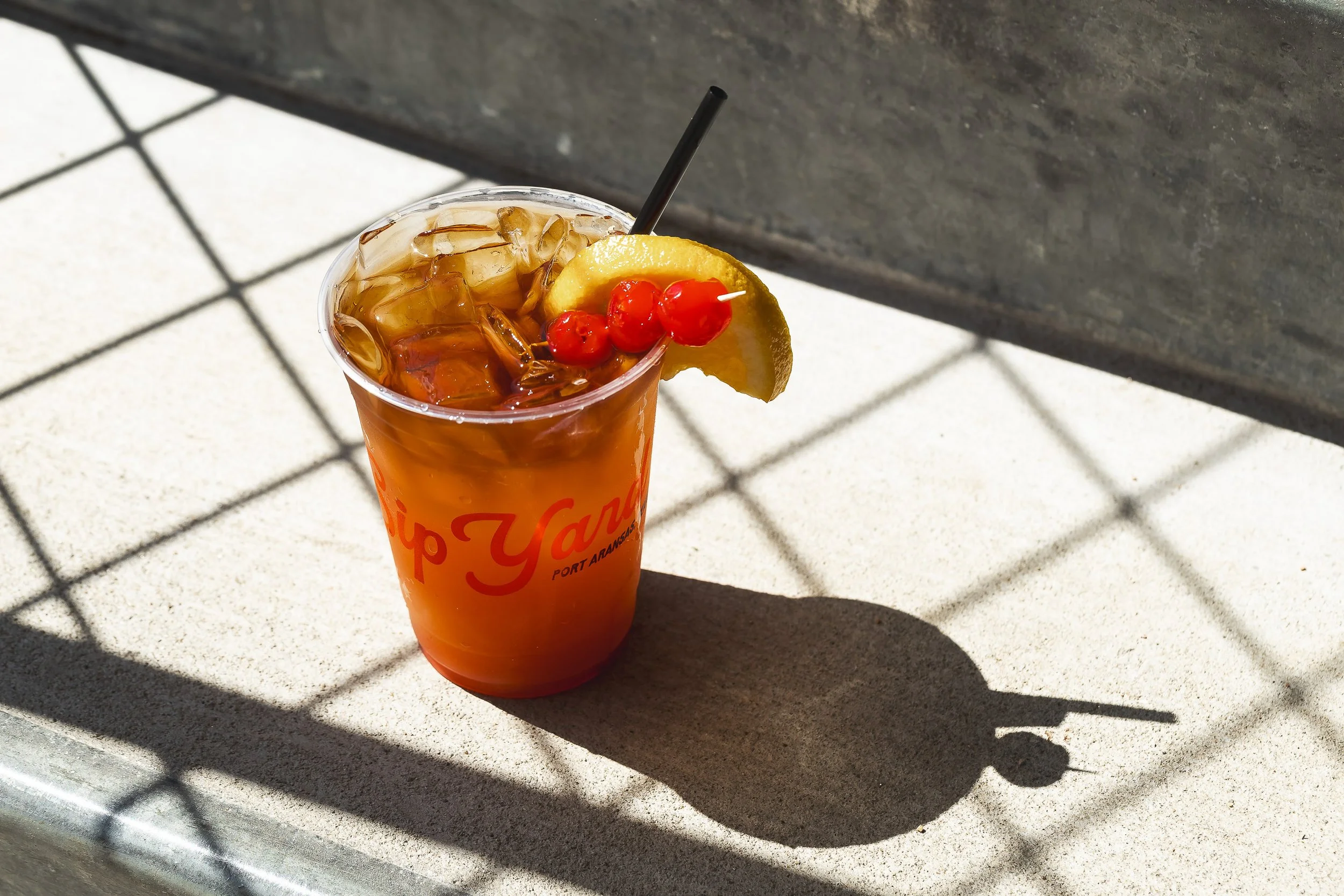 Iced amber cocktail in branded plastic cup with lemon wedge and cherries casting strong outdoor shadow
