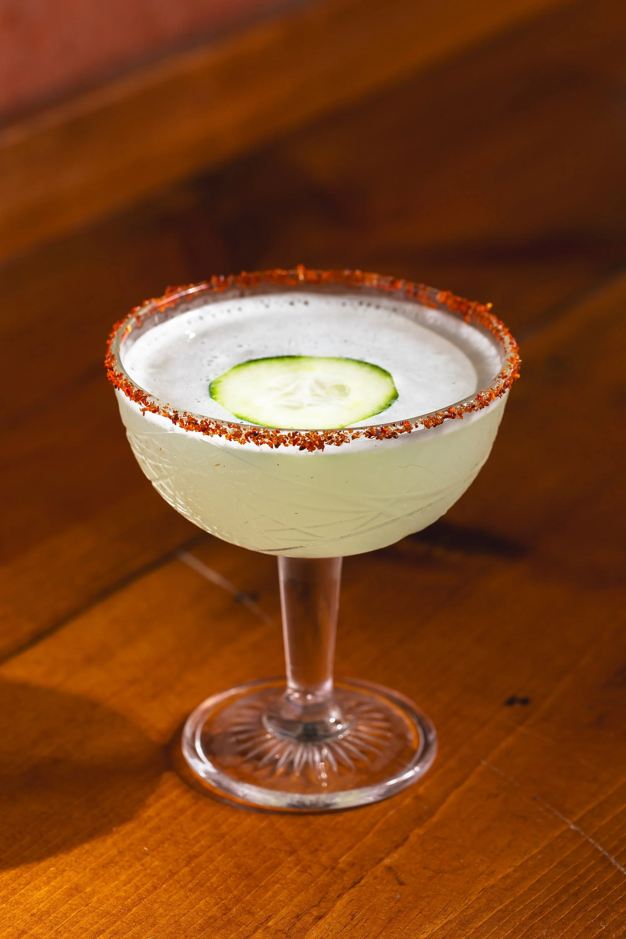 Pale green craft cocktail in etched coupe glass with chili-salt rim, cucumber garnish, and warm wood bar backdrop
