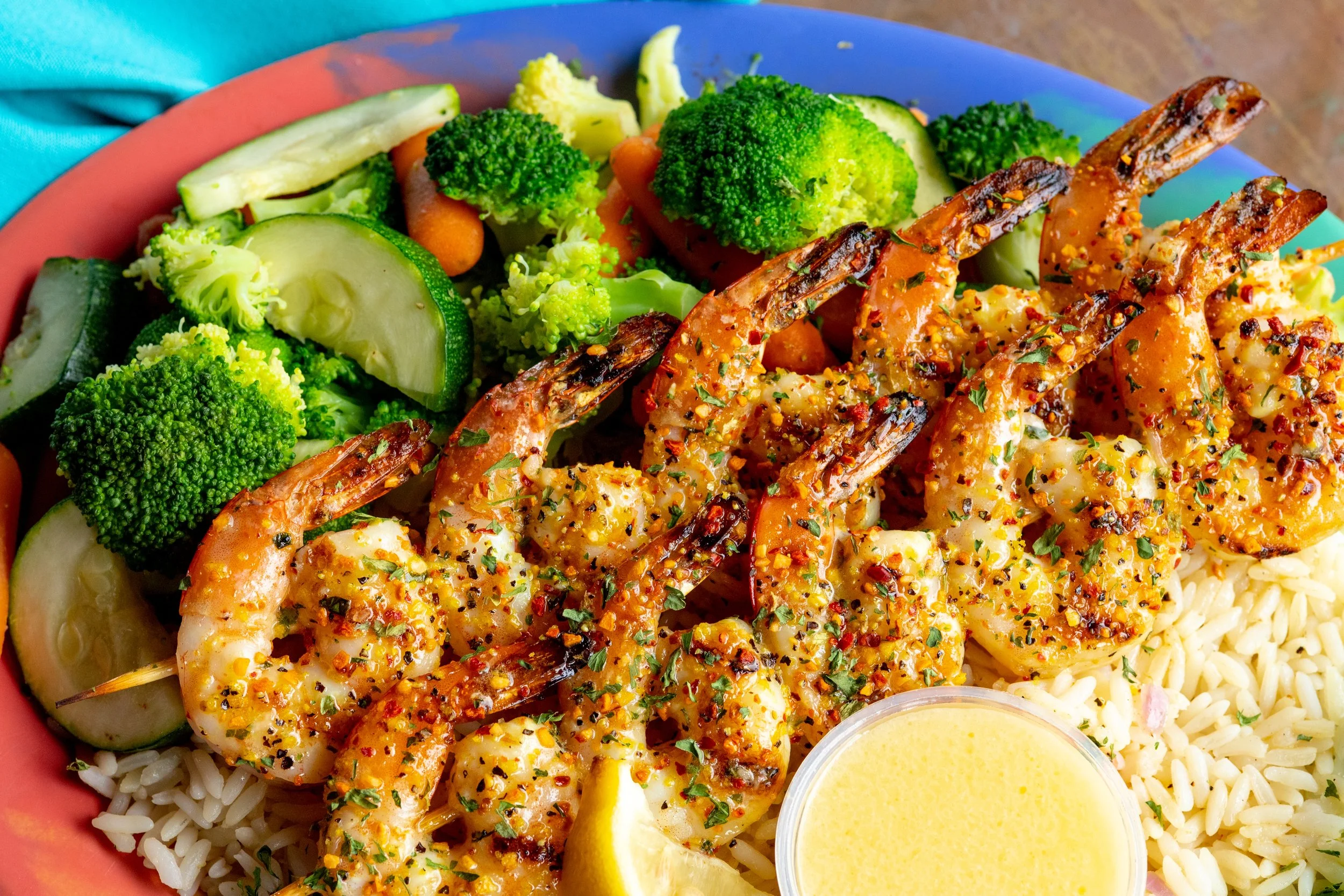 Grilled shrimp seasoned with spices, served over white rice with steamed broccoli, zucchini, carrots, and a lemon butter sauce on a colorful plate.