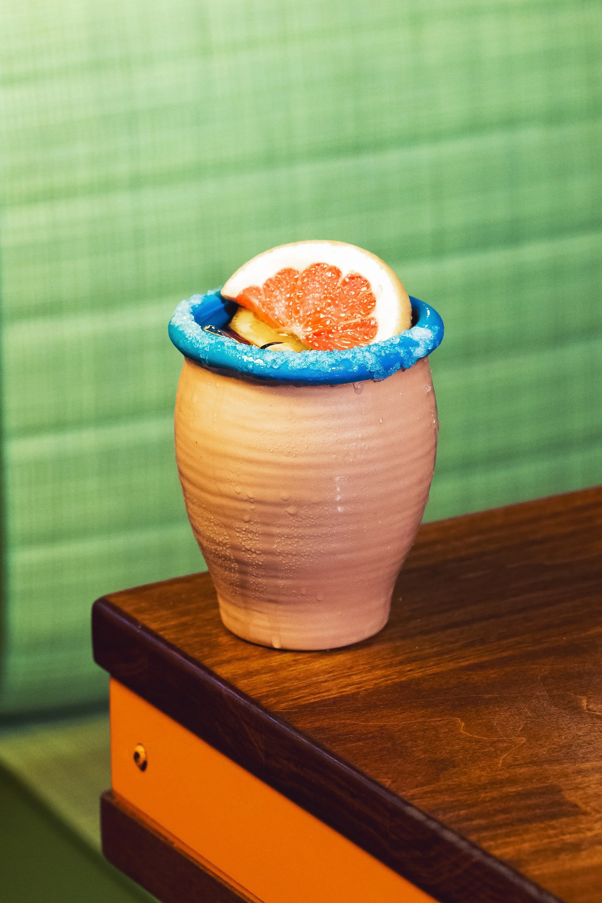 Pink grapefruit cocktail in ceramic cup with blue sugar rim and citrus garnish on restaurant table
