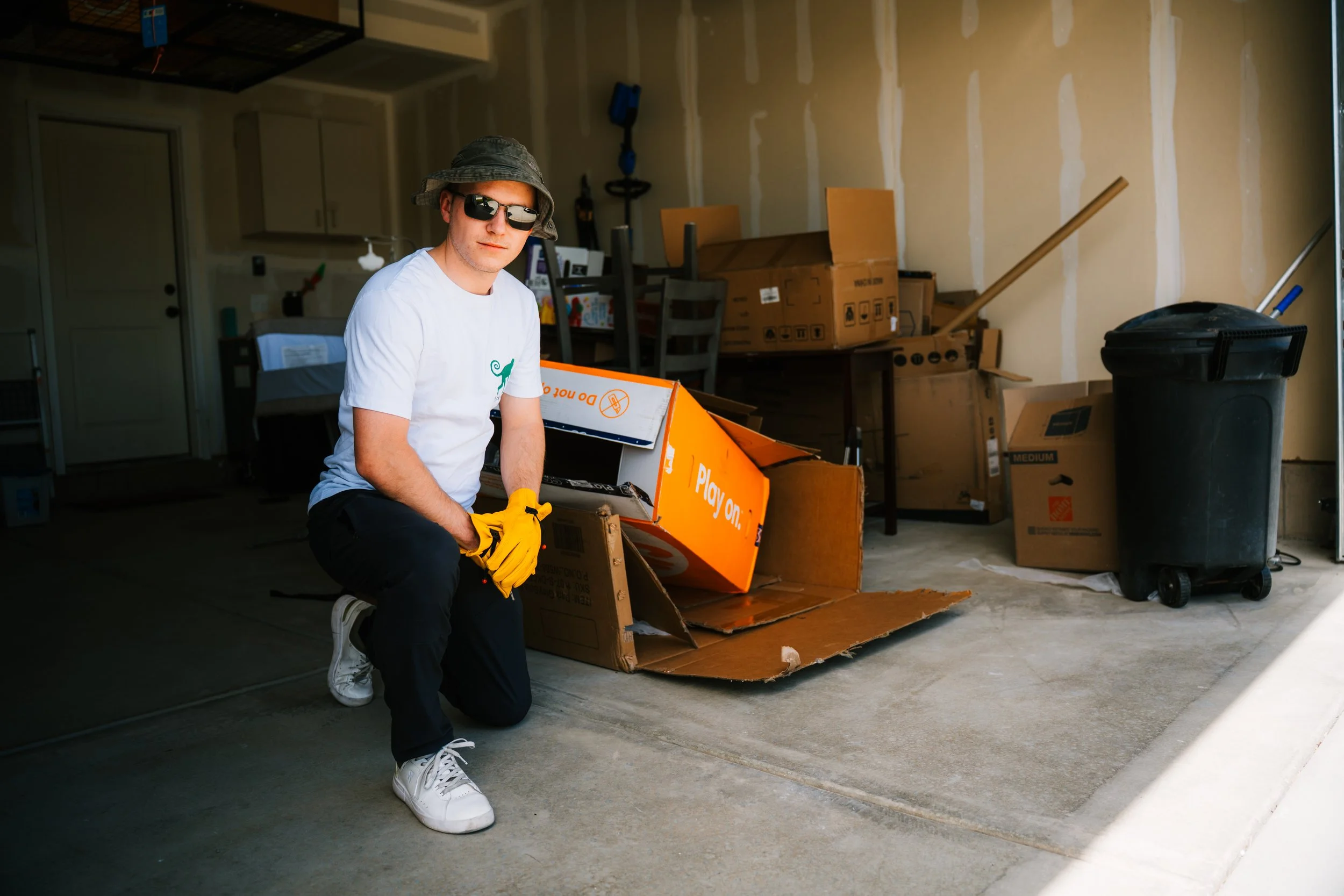 A young man kneels on the concrete driveway in a garage, wearing sunglasses, a bucket hat, a white t-shirt, and yellow gloves. He is holding a tool and surrounded by opened and crushed cardboard boxes, with some stored or discarded items around, including a large trash bin.