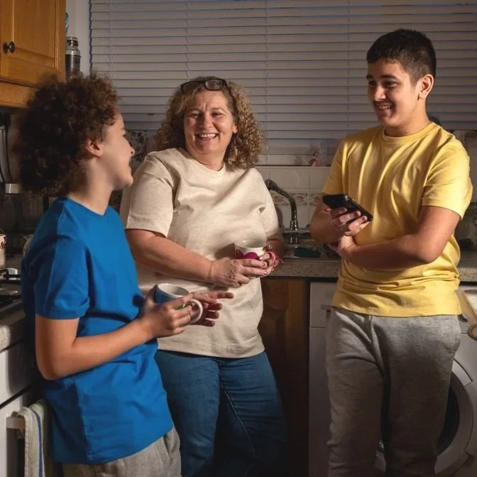 A woman and two boys talk and laugh in a kitchen, holding mugs and a phone, during a casual gathering.