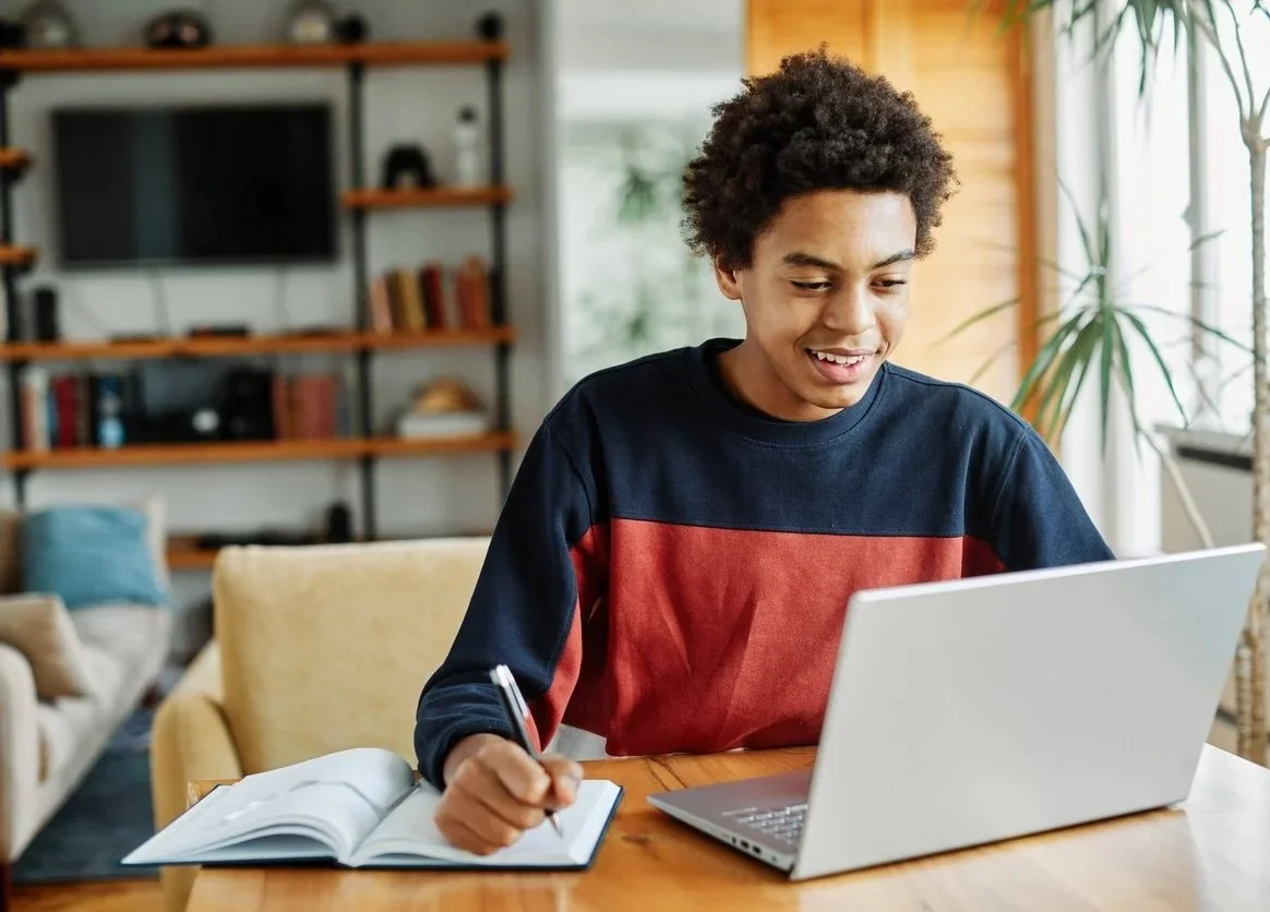 A teenage boy with curly hair using a laptop and writing in a notebook at a wooden table in a living room.