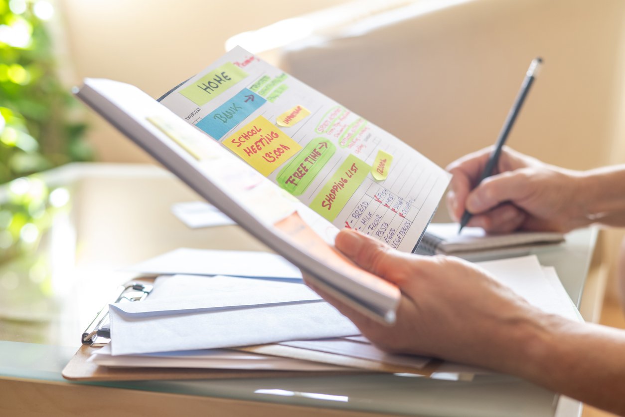 Person organizing a colorful handwritten weekly planner or journal on a desk with papers and a clipboard nearby, with sunlight streaming through a window.