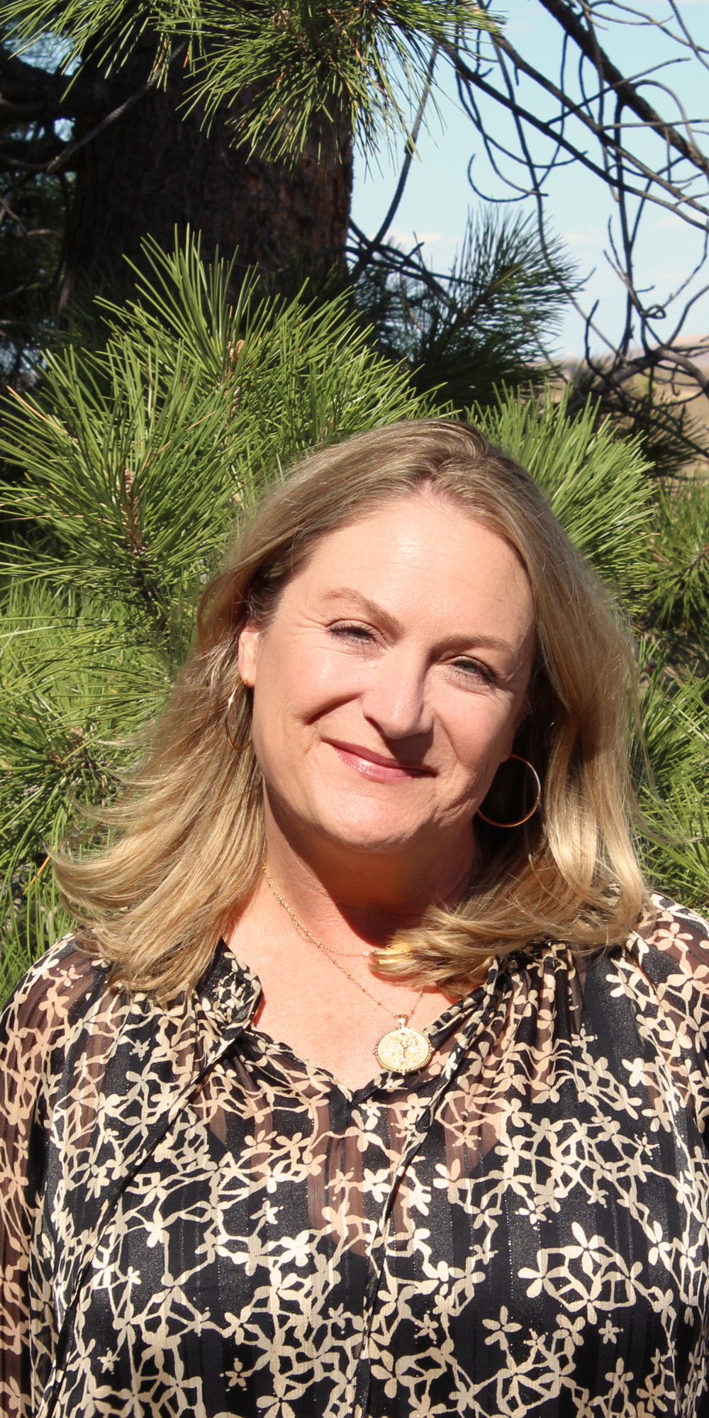 Lisa Kennedy smiling outdoors, in front of a green pine tree