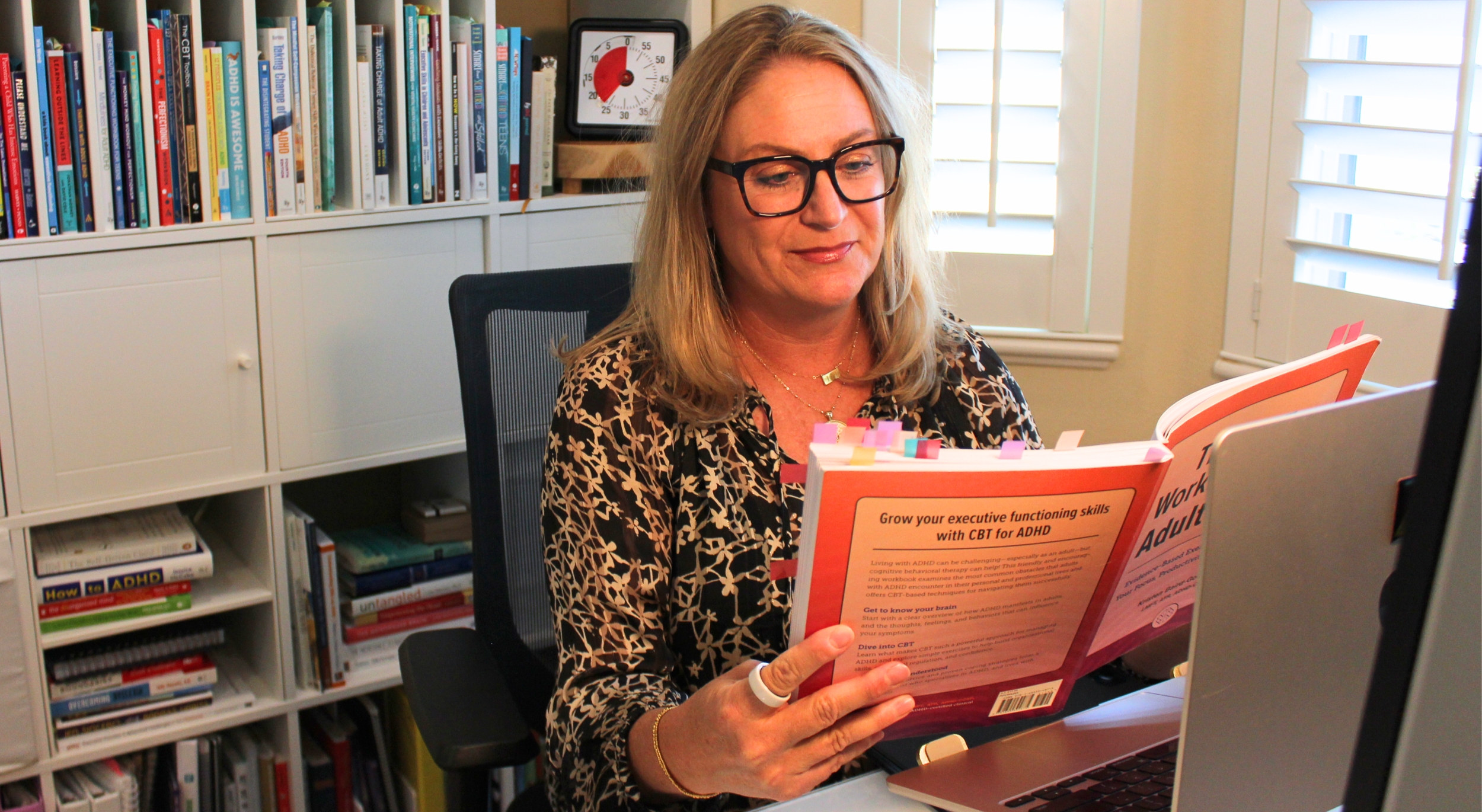 Lisa wearing glasses and sitting at a desk reading a book about executive functioning skills, with bookshelves filled with books in the background