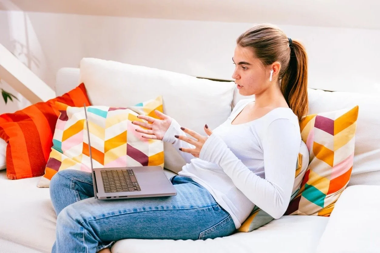 Young woman sitting on a white couch with colorful geometric pillows, using a laptop and gesturing with her hands, wearing white long-sleeve shirt and wireless earbuds.