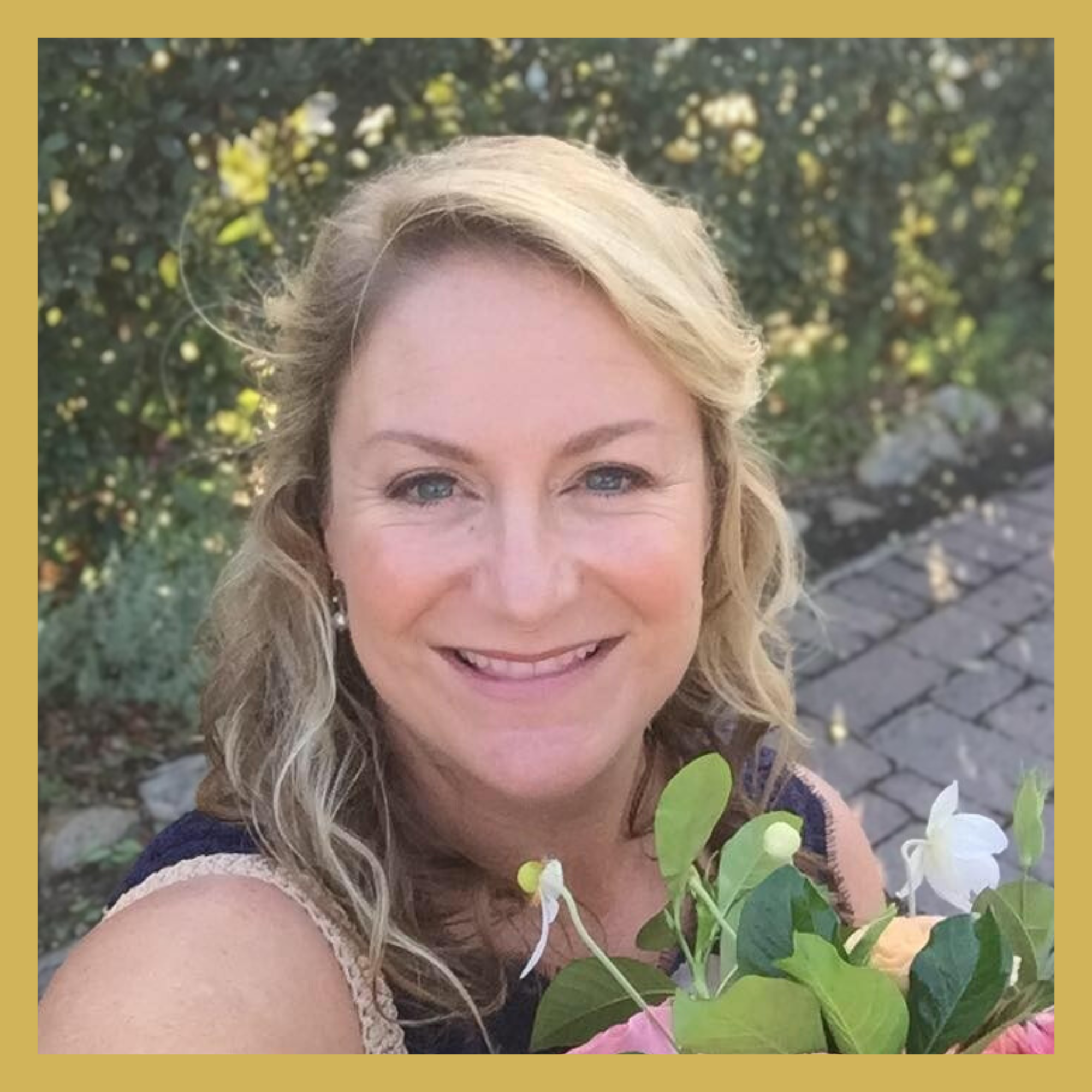 A woman with blonde curly hair smiling outdoors surrounded by green leaves and colorful flowers.