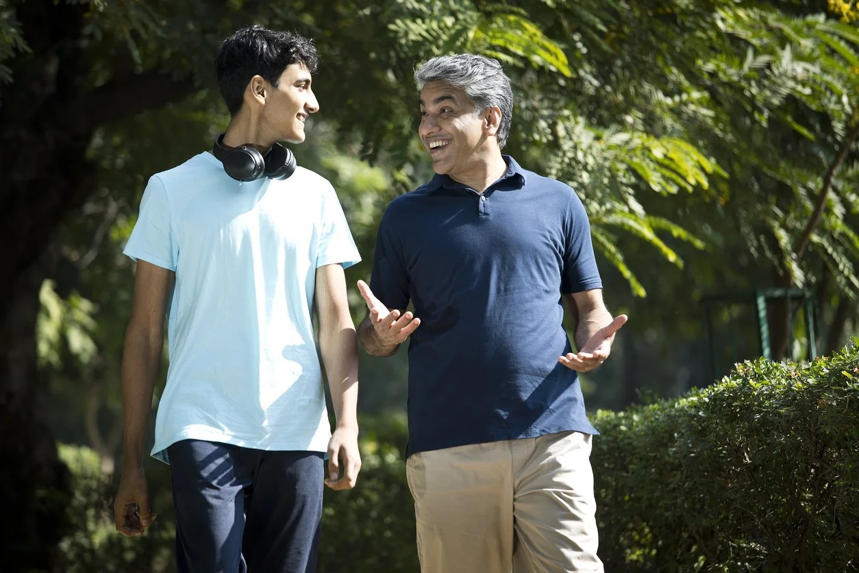 A young man and an older man are walking and talking outdoors in a park, with trees and greenery around them. The older man is smiling and gesturing with his hands, while the young man, wearing headphones around his neck, listens and smiles.