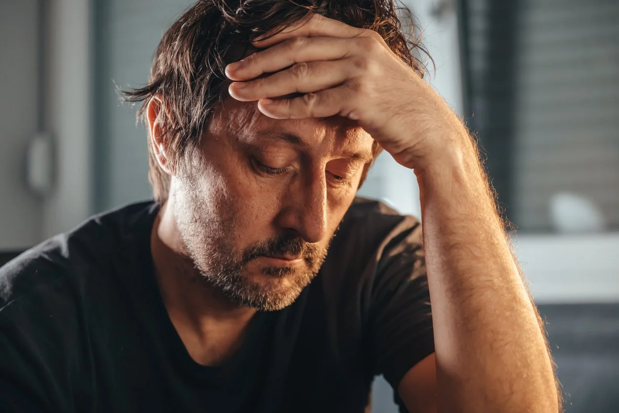 Man with brown hair and beard holding his head and looking down in distress or frustration.