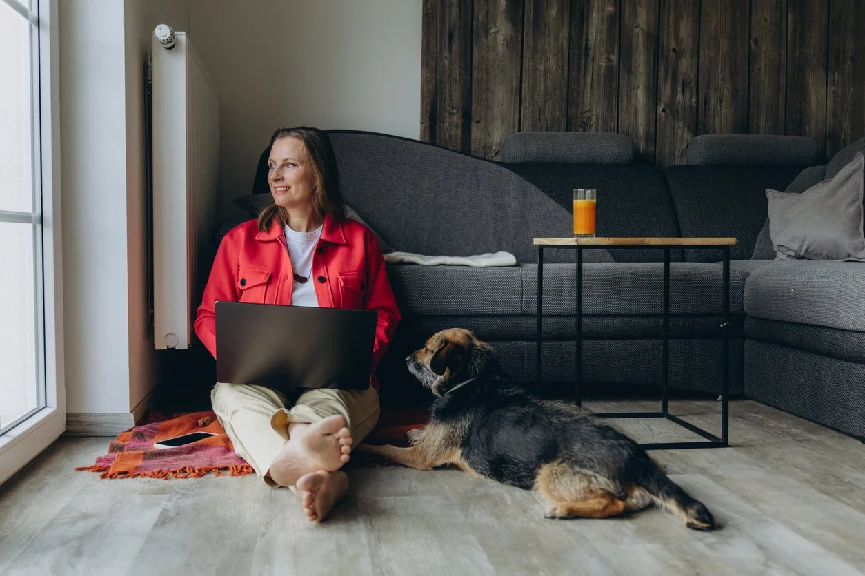 A woman sitting on the floor with a laptop, next to a dog, in a cozy living room with a glass of orange juice on a side table.