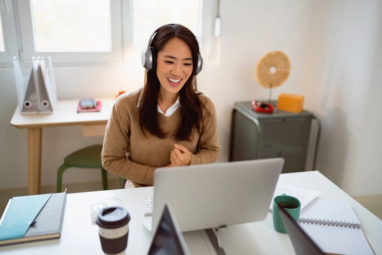 A woman with long dark hair wearing headphones, smiling and looking at her laptop in a bright, cozy home office.