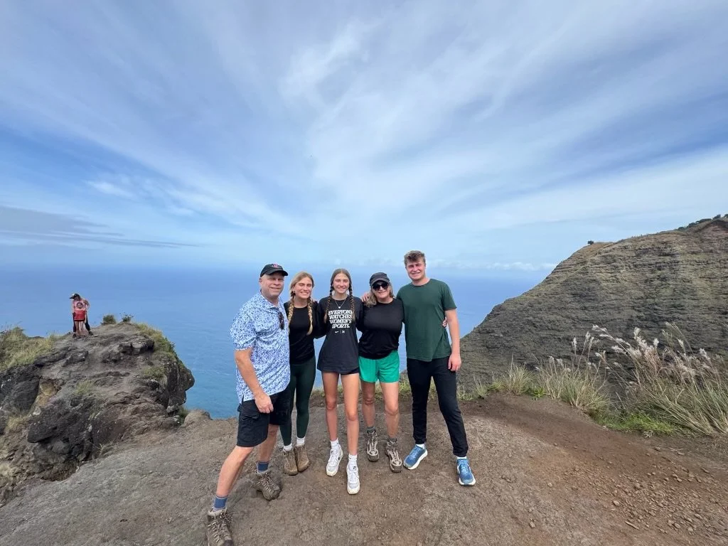 Lisa's family standing together on a cliff overlooking the ocean, with a large hill on the right and cloudy sky above, posing for a photo.