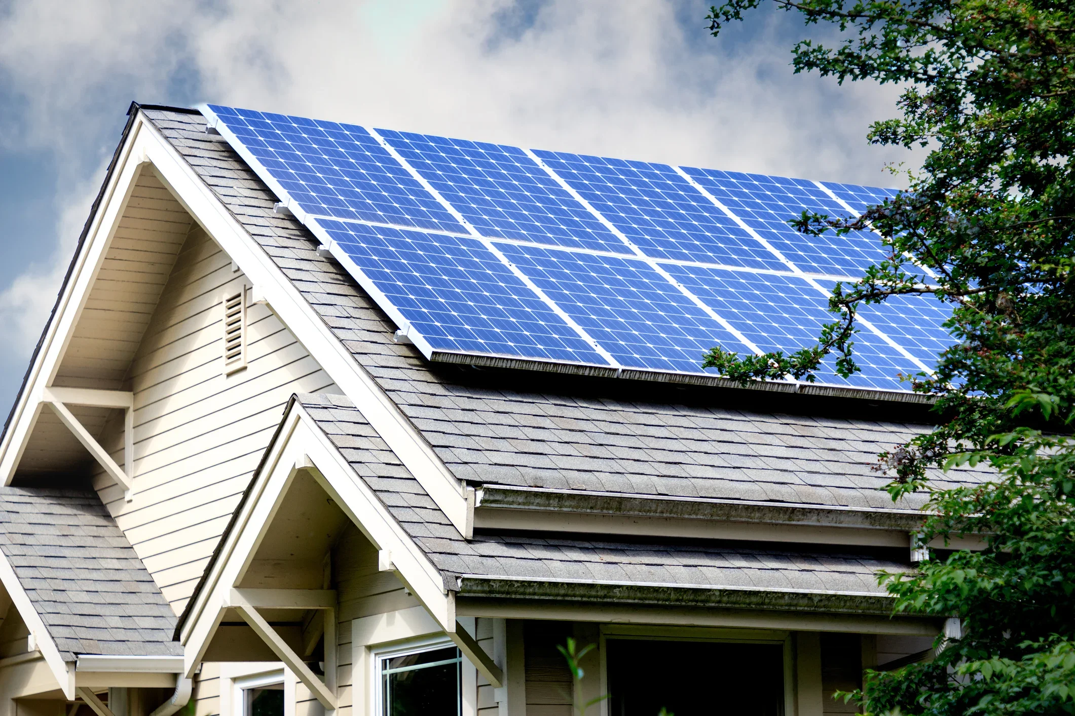 A house with solar panels on the roof and gray shingled roof, surrounded by green trees and partly cloudy sky.