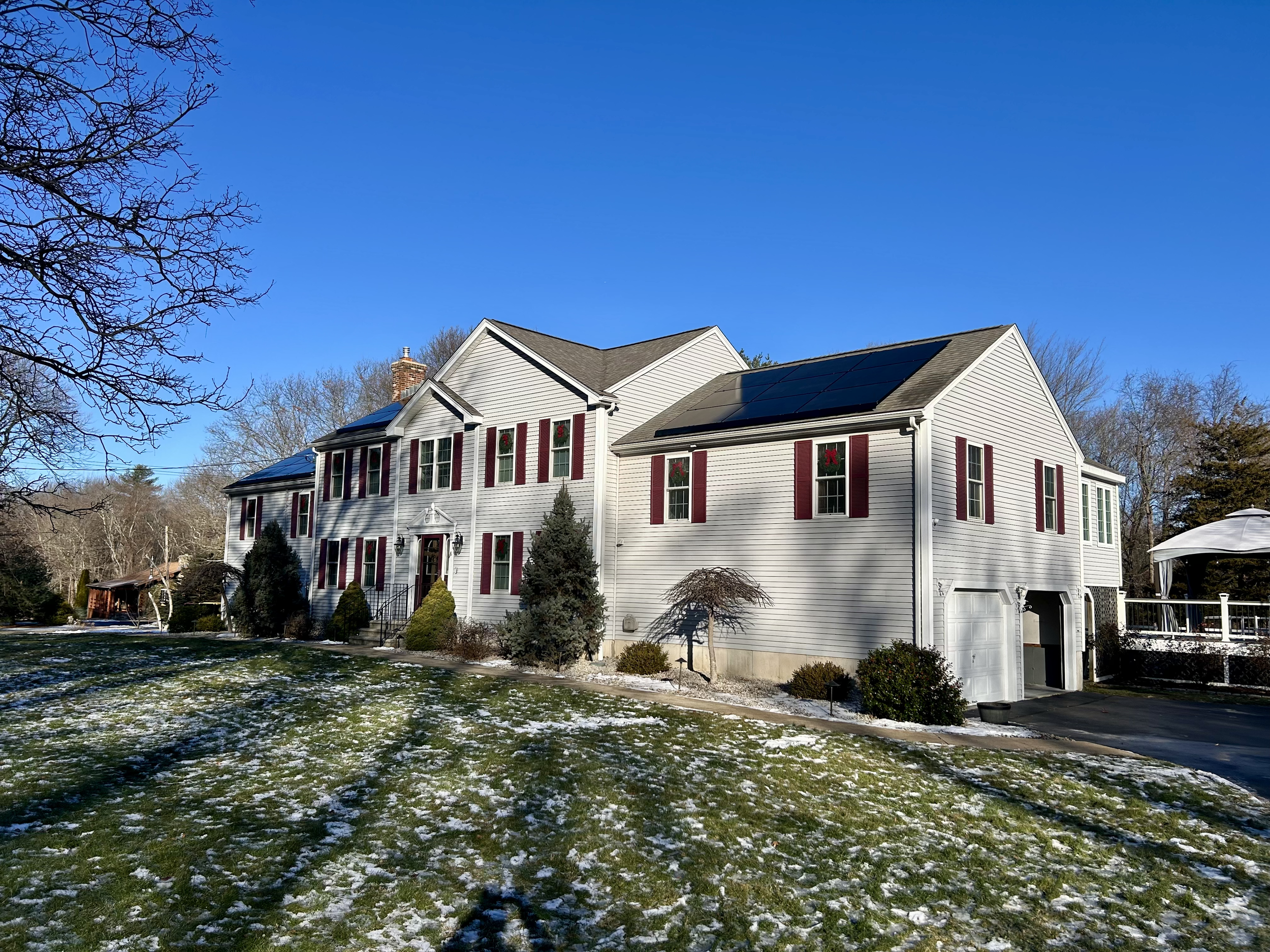 A large white house with red shutters and solar panels on the roof, decorated for Christmas with wreaths, on a snowy lawn under a clear blue sky.