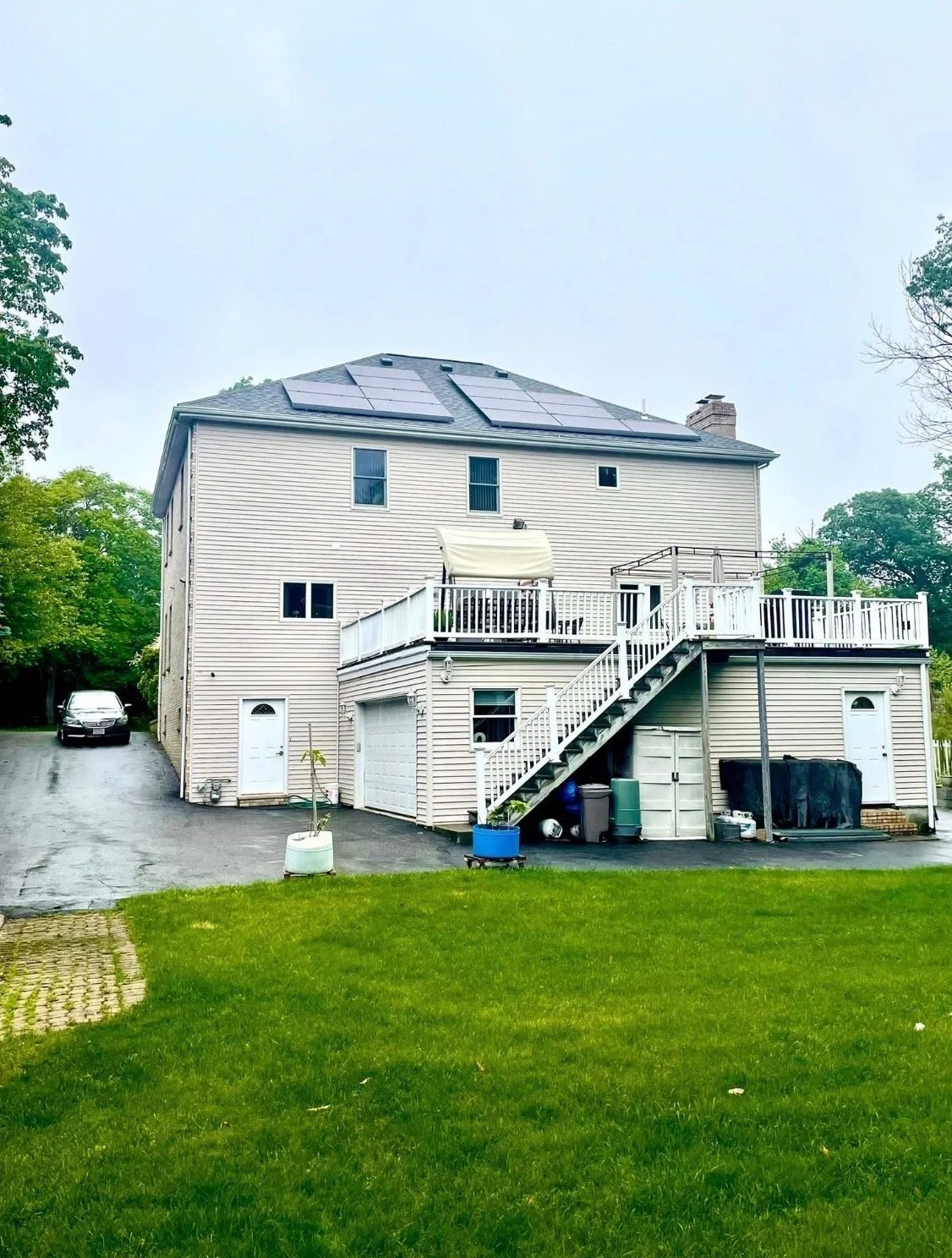 A multi-story house with solar panels on the roof, a large deck with outdoor furniture, and a staircase leading down to a backyard lawn.
