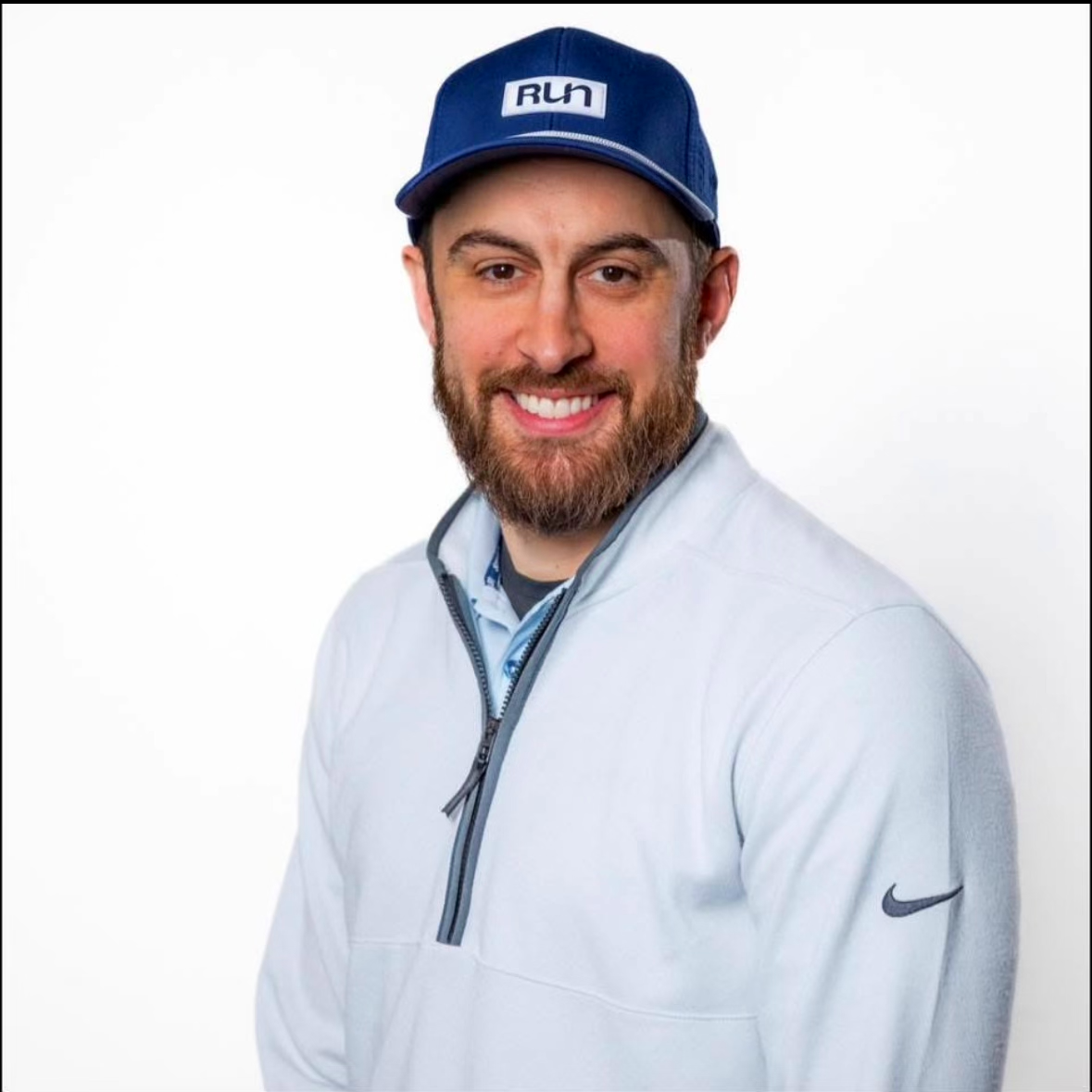 A smiling man with a beard wearing a blue cap with the word "Run" on it, a white jacket, and a collared shirt.