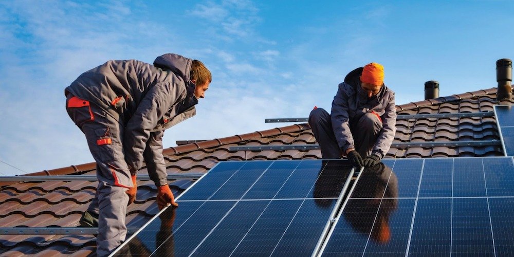 Two workers install solar panels on a tiled roof under a blue sky.