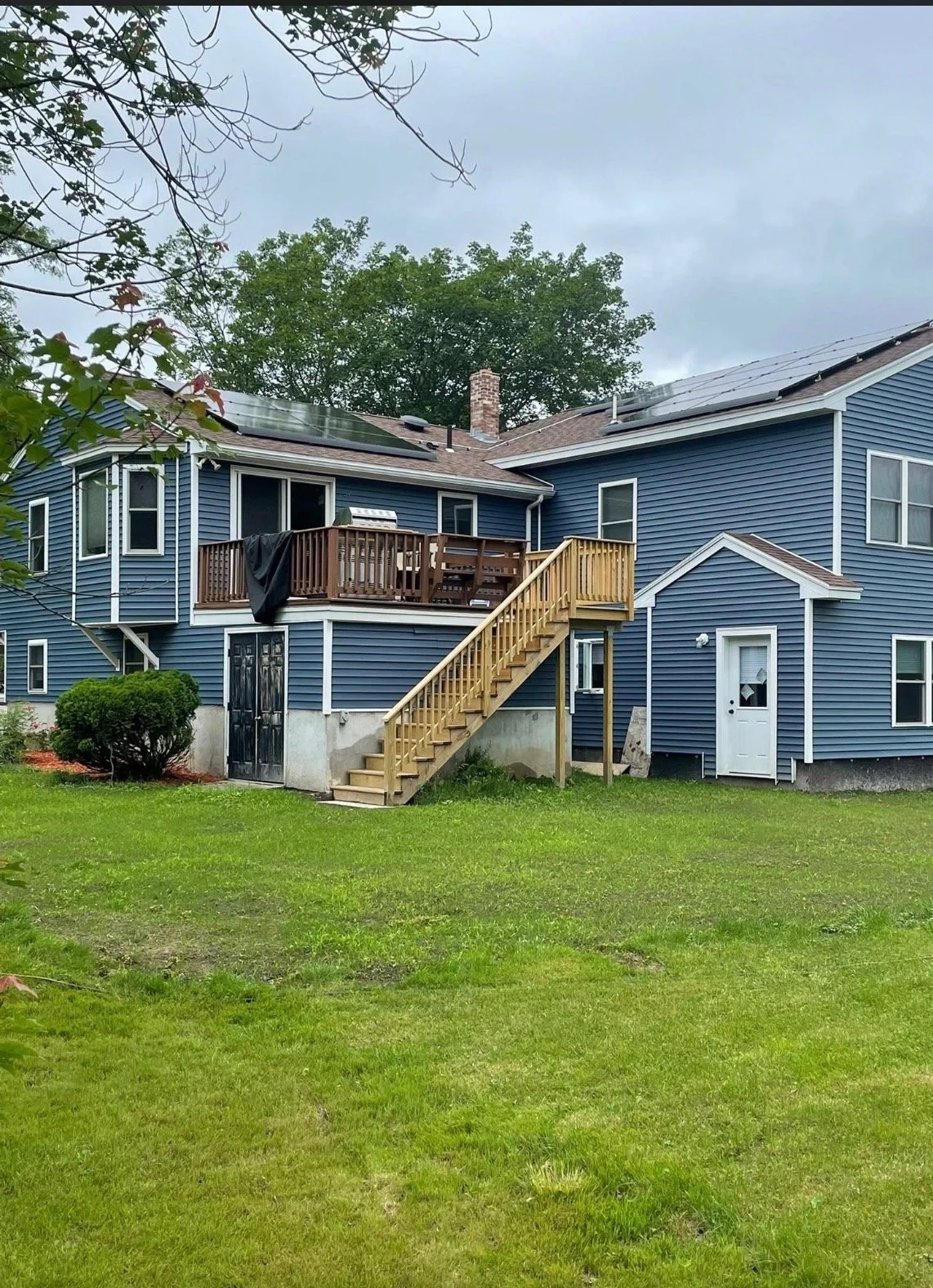 A blue two-story house with a wooden staircase leading to an upper deck, which has outdoor furniture and a black cloth hanging on the railing. The house has a small white door on the lower right and multiple windows. The yard is grassy with a small bush and some trees, under an overcast sky.
