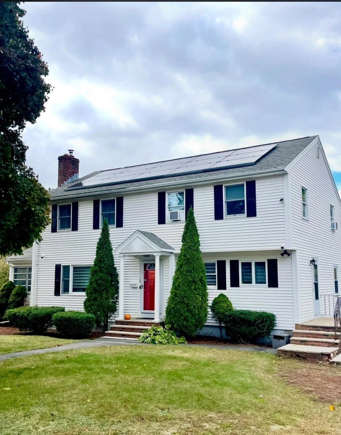 A two-story white house with black shutters, a red front door, and a small porch with steps. The house has solar panels on the roof and landscaping with bushes and tall, narrow trees in the front yard. There is a pumpkin on the steps and a cloudy sky overhead.