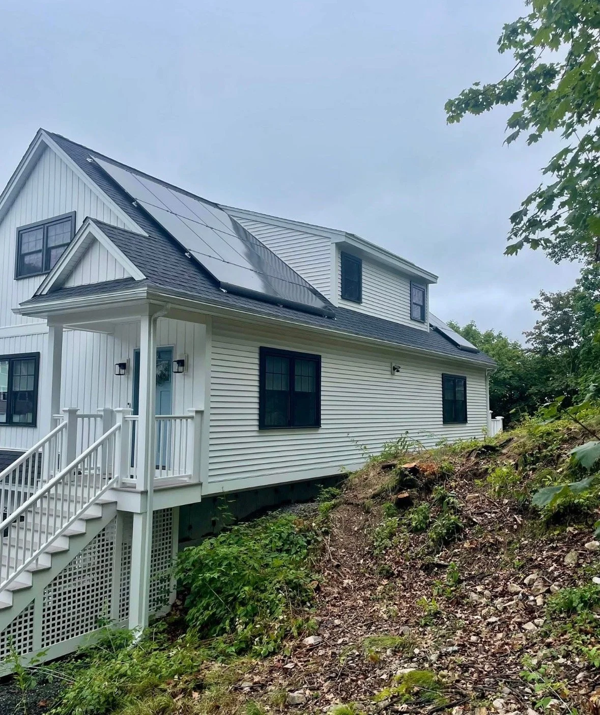 A white two-story house with black window frames and solar panels on the roof, with a small front porch and stairs, situated on a slightly sloped terrain with bushes and soil in the foreground.