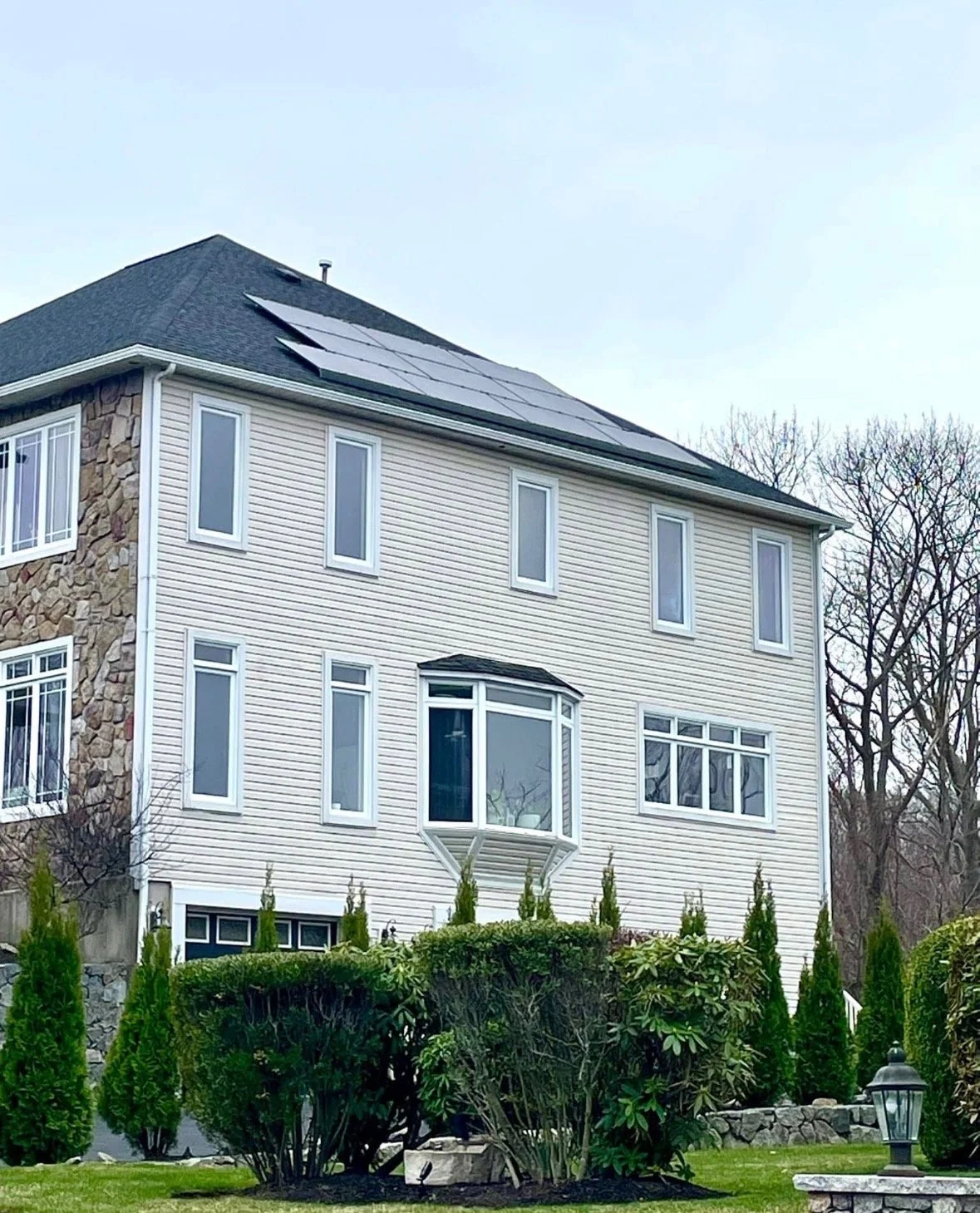 A multi-story house featuring a stone and vinyl siding exterior, with a roof equipped with solar panels, multiple windows including a prominent bay window, and landscaped bushes in the front yard.